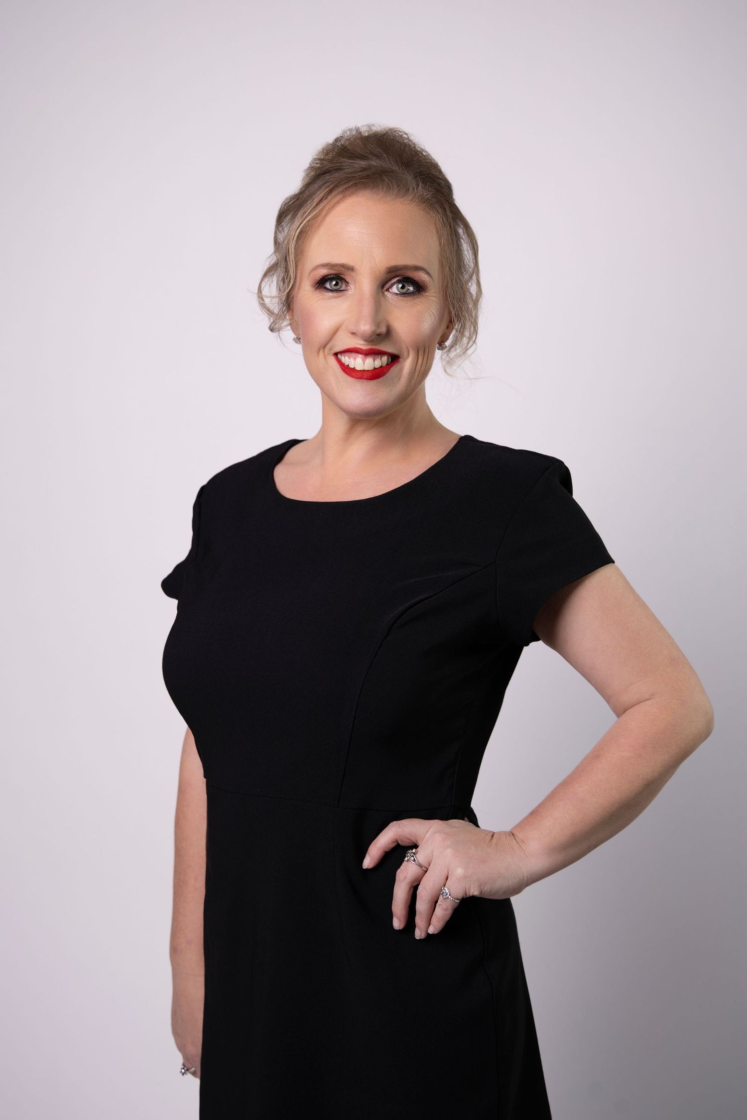 Blonde woman in black dress with red lipstick posing confidently against a white studio background.