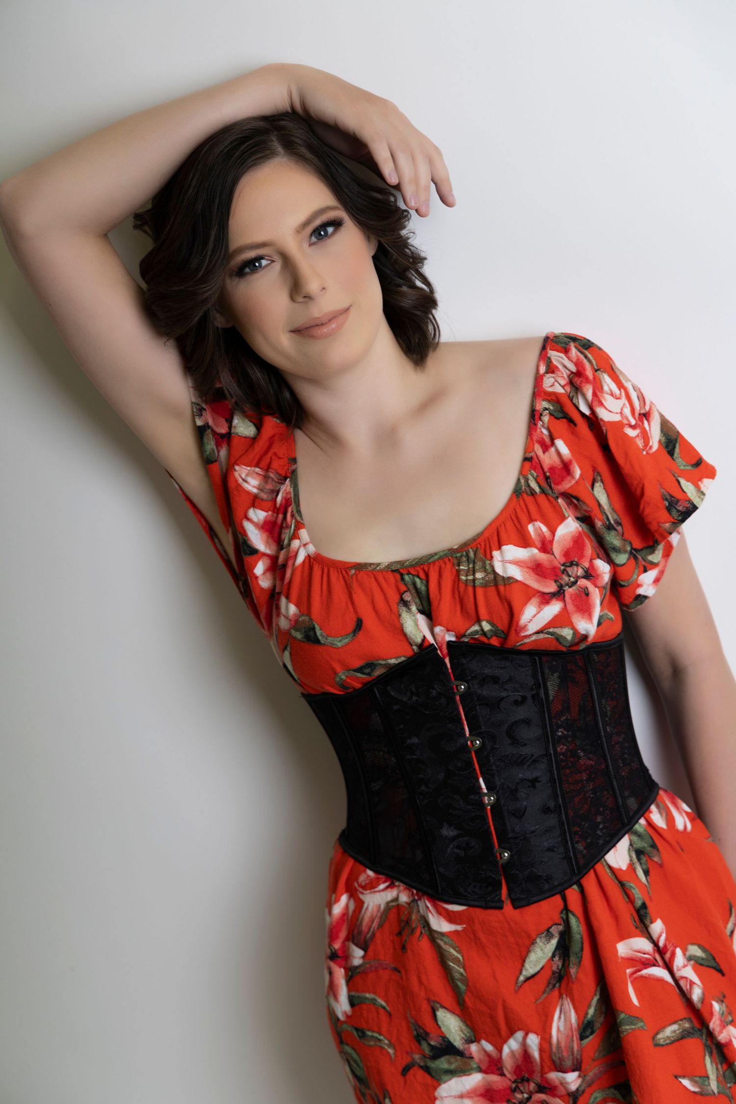 Woman in red floral dress with black corset posing against white wall.