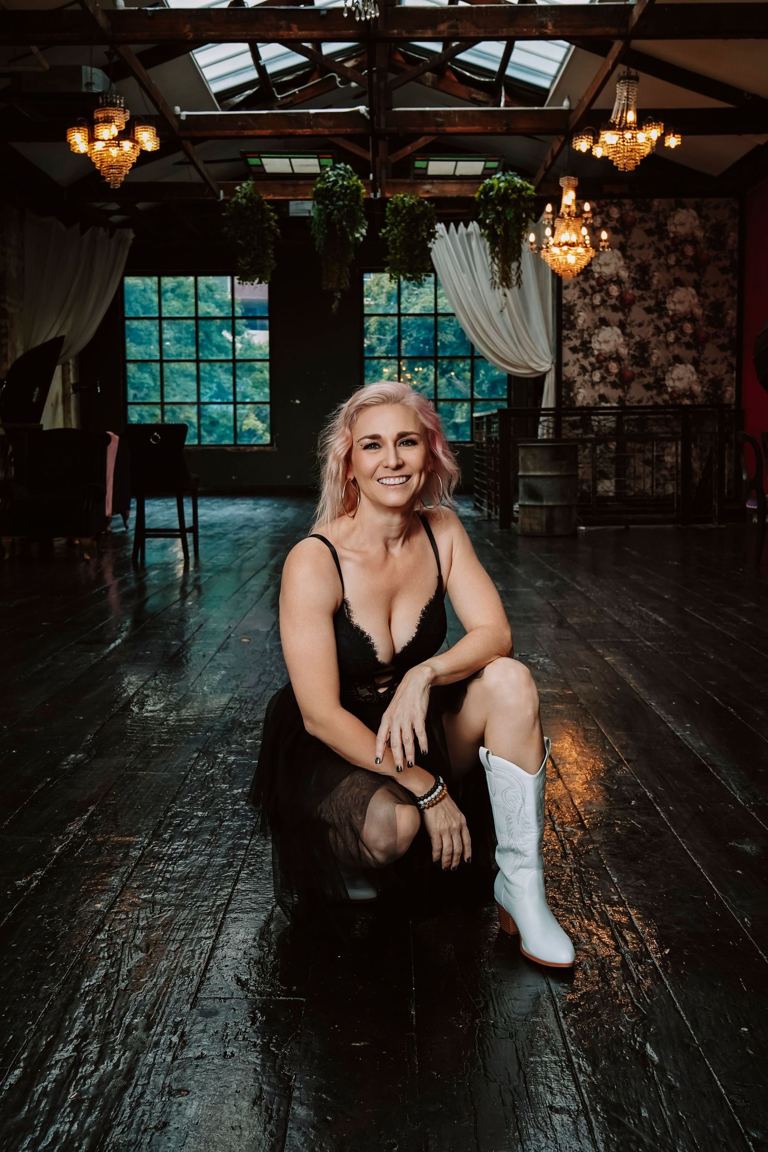 Smiling woman with pink hair in black dress and white boots sitting on dark floor.
