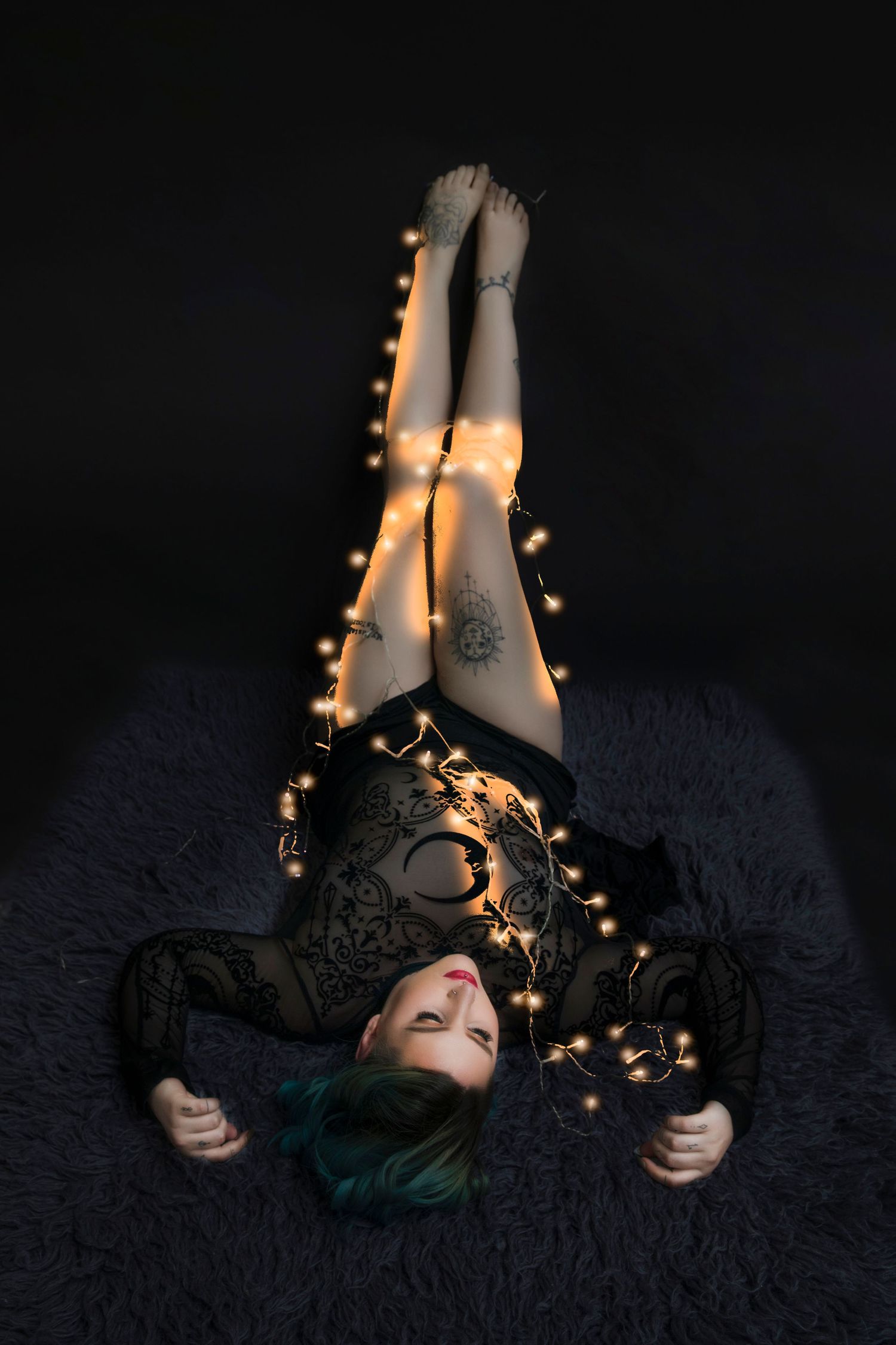 Woman with teal hair lying on dark fur with legs raised, wrapped in glowing string lights.