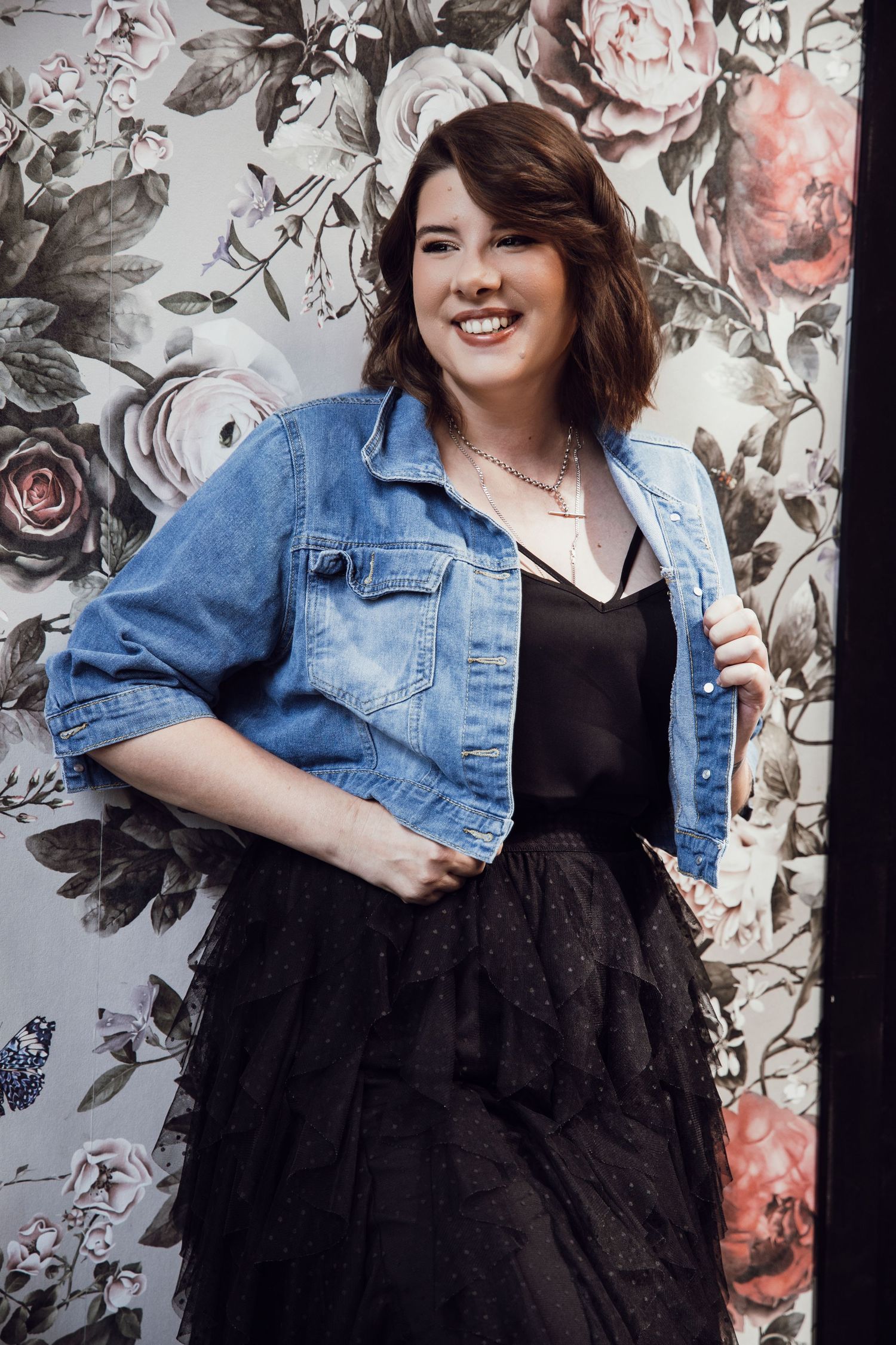 Smiling woman in denim jacket and black tulle skirt poses against floral wallpaper.