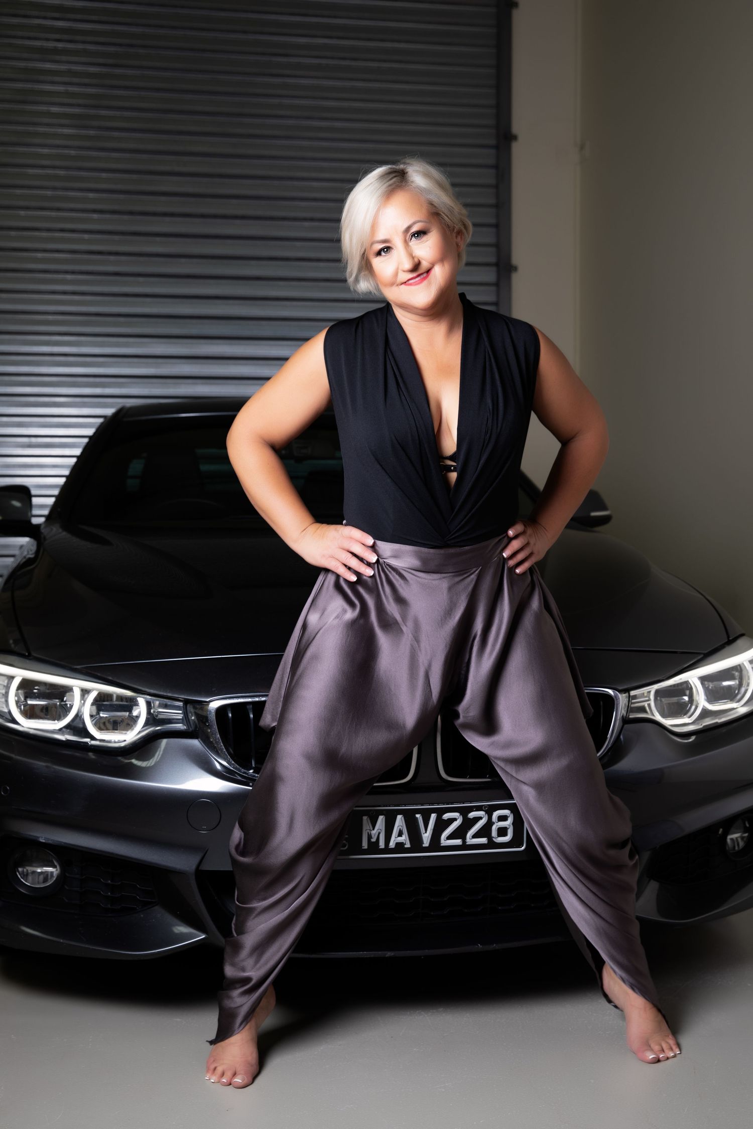 Confident blonde woman in black top and gray pants posing in front of dark BMW car.