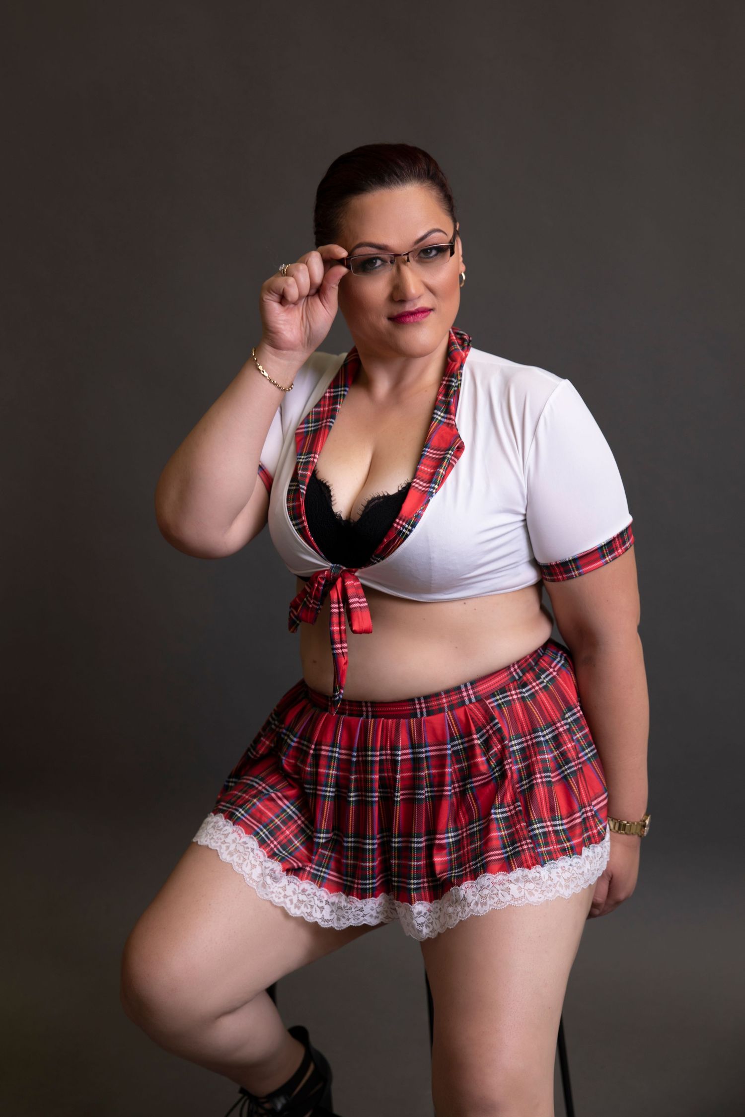 Plus size woman in red plaid schoolgirl skirt and white crop top posing with glasses.