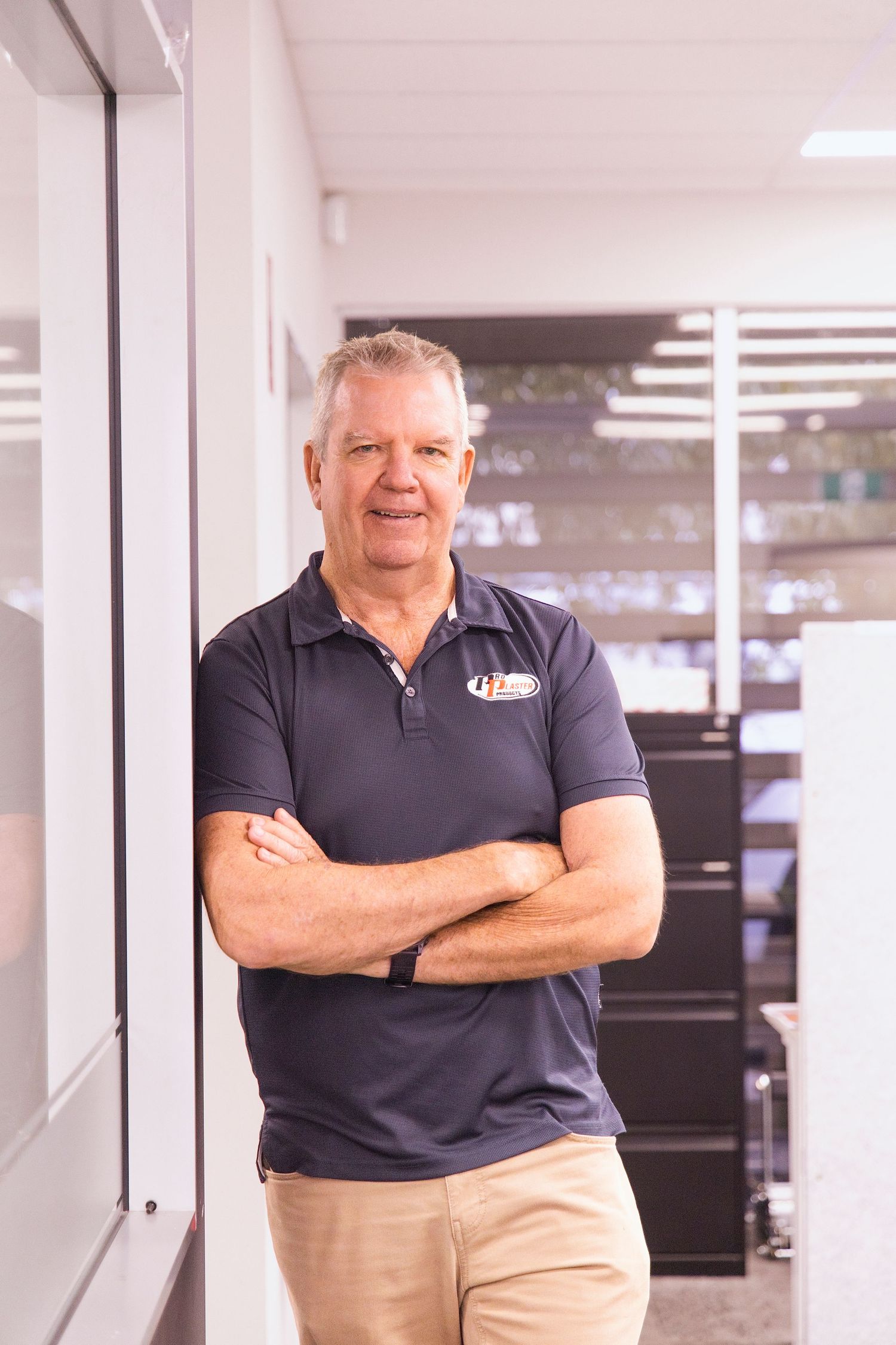 Older man in navy polo shirt stands arms crossed leaning against wall in modern office hallway.