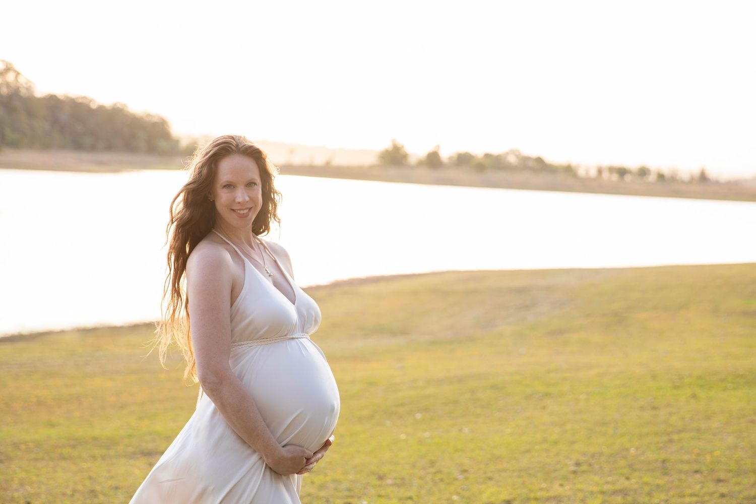Smiling pregnant woman in white dress holding baby bump near lakeside in warm golden light.