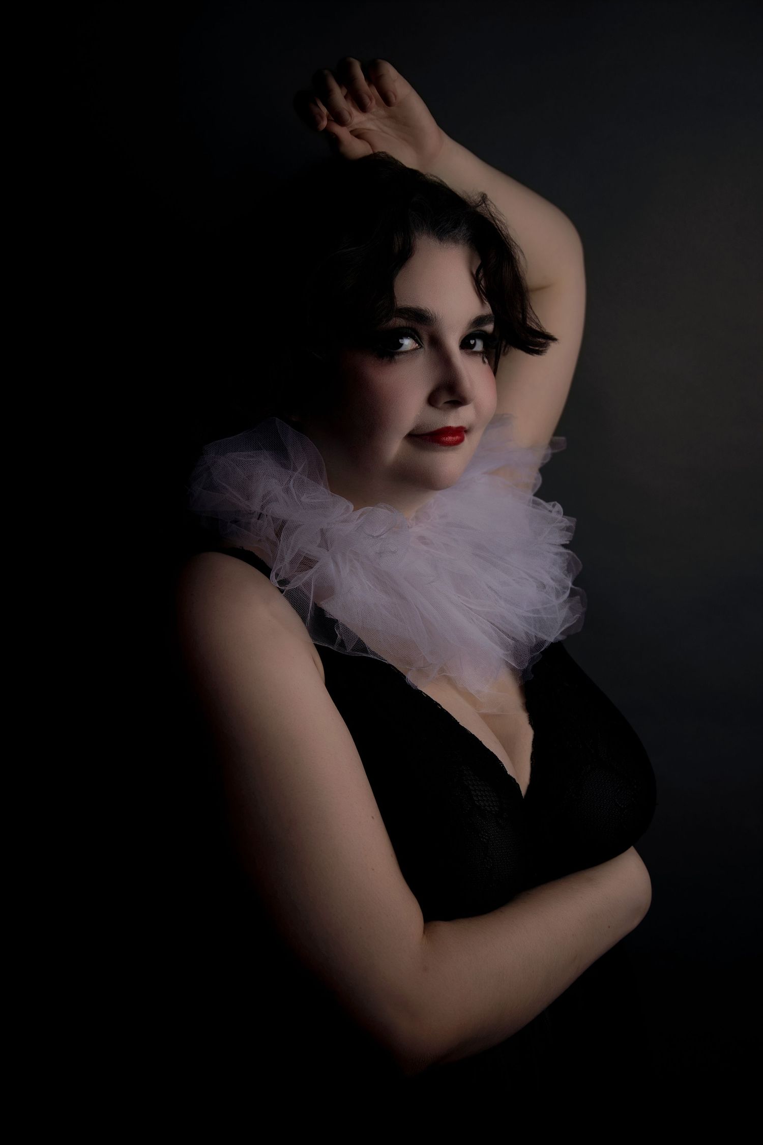Dramatic dark portrait of woman in black dress with white ruffled collar and bold red lipstick.