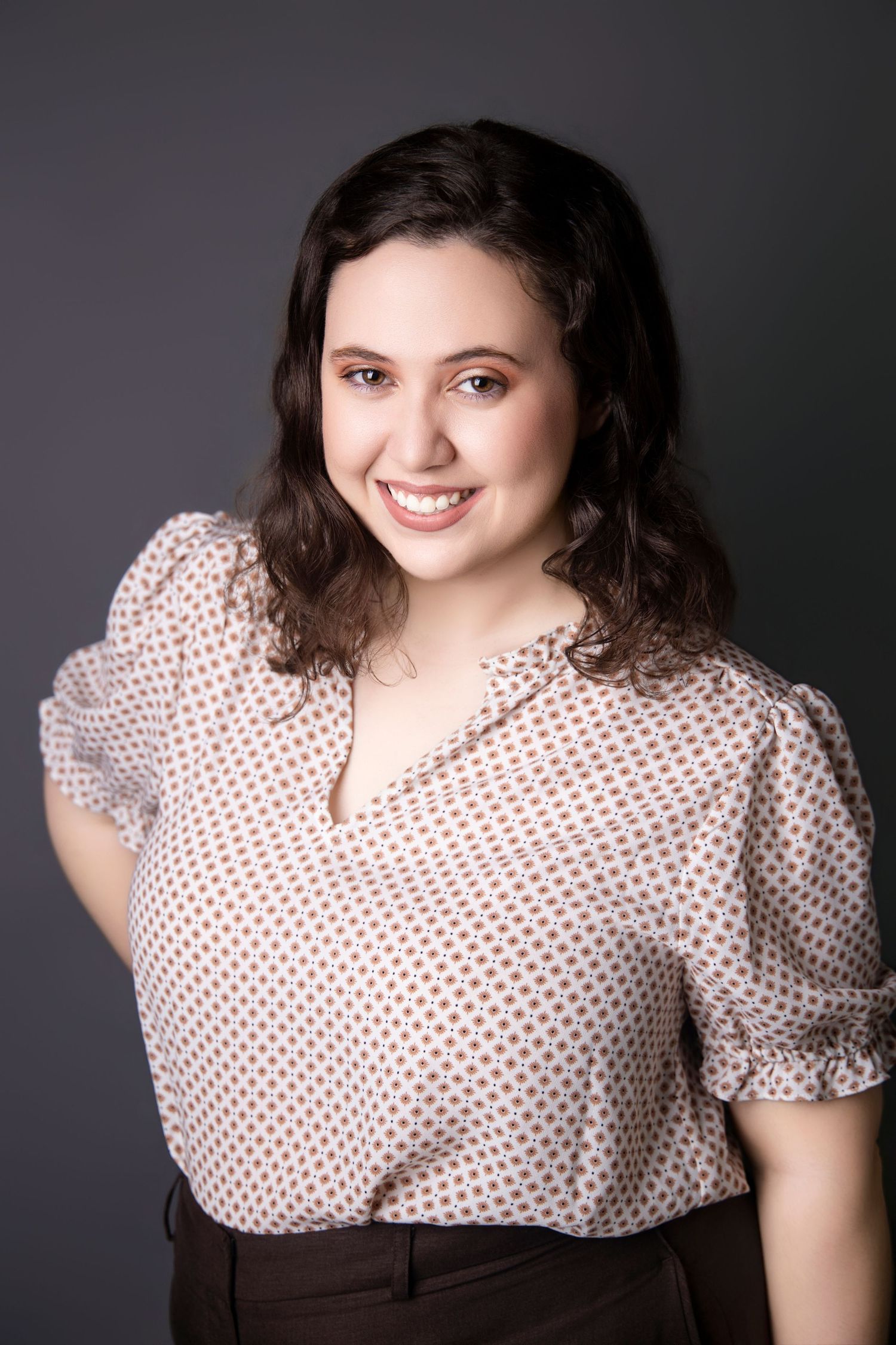 Young brunette woman in polka dot blouse smiling confidently against dark gray studio backdrop.