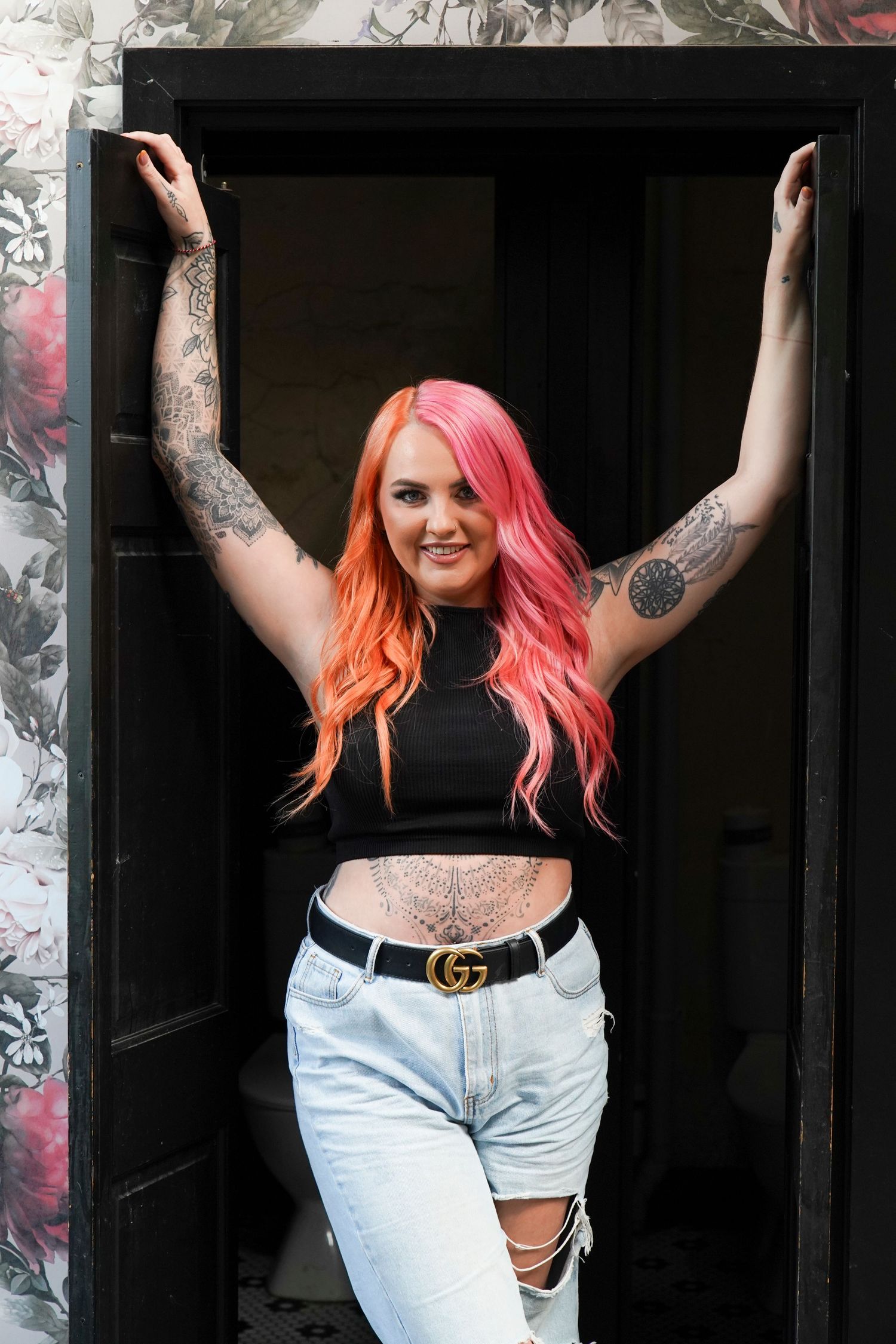 Woman with pink orange hair and tattoos in black crop top and jeans in doorway.