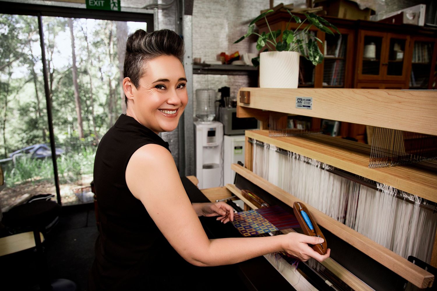 Woman smiling at camera while working at a wooden loom in a bright studio with green trees outside.