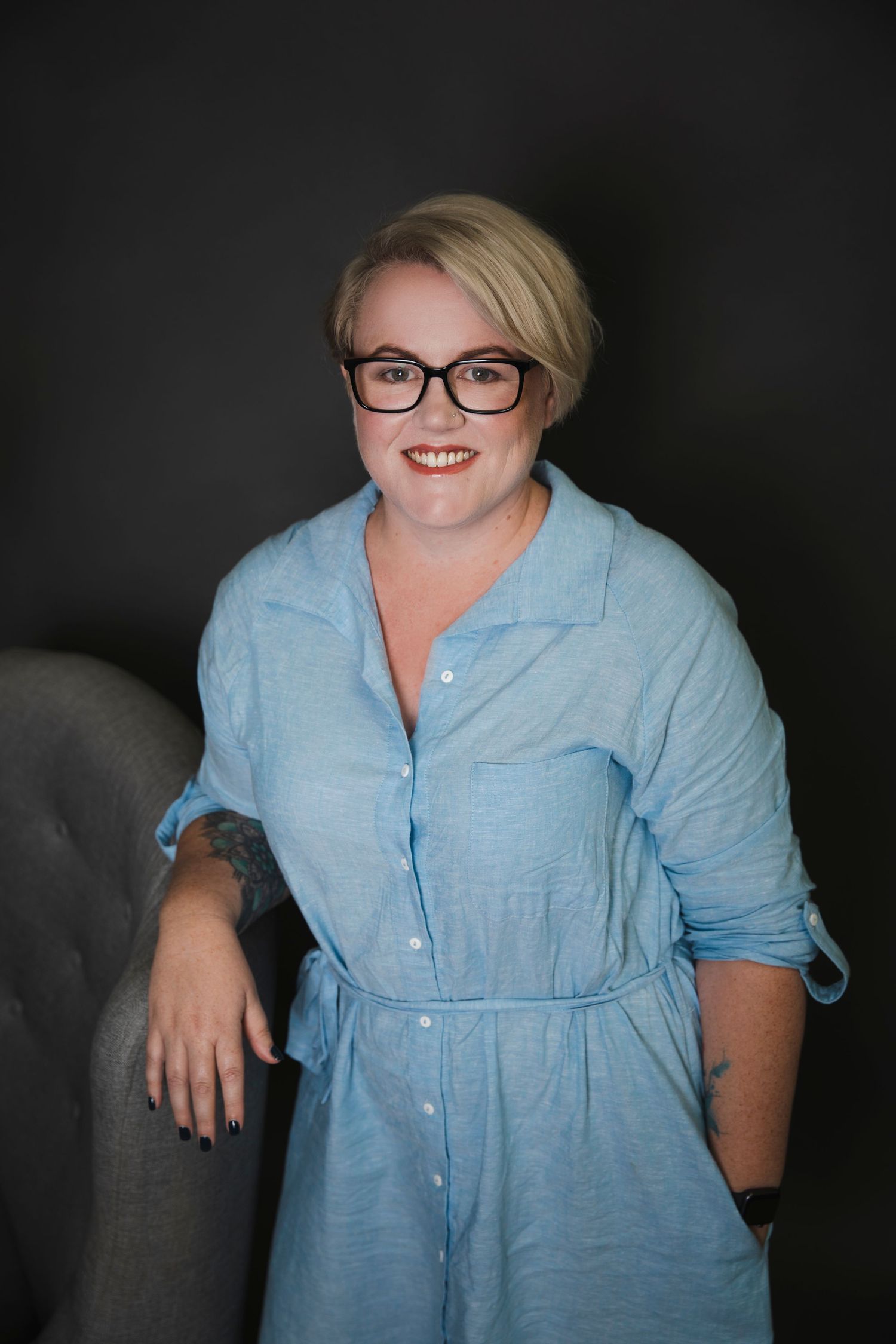 Smiling blonde woman with glasses in blue dress leaning on chair in dark studio.