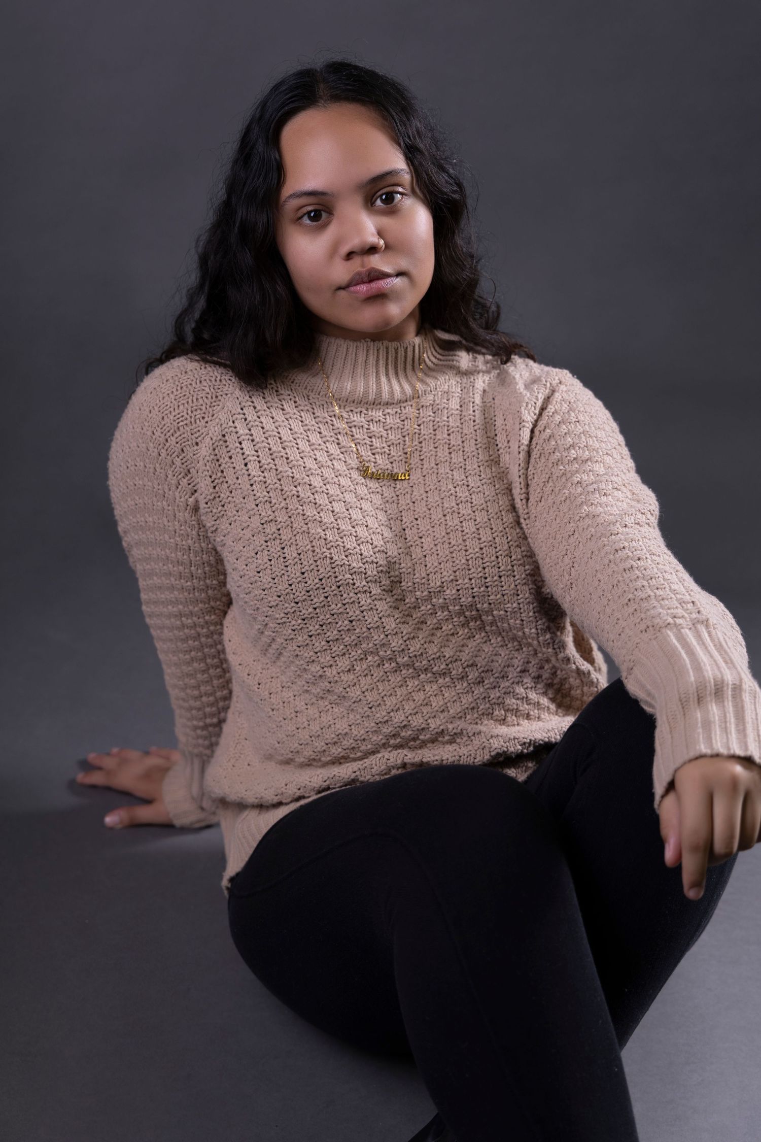 Young woman in beige knit turtleneck sweater and black pants sitting in studio portrait.