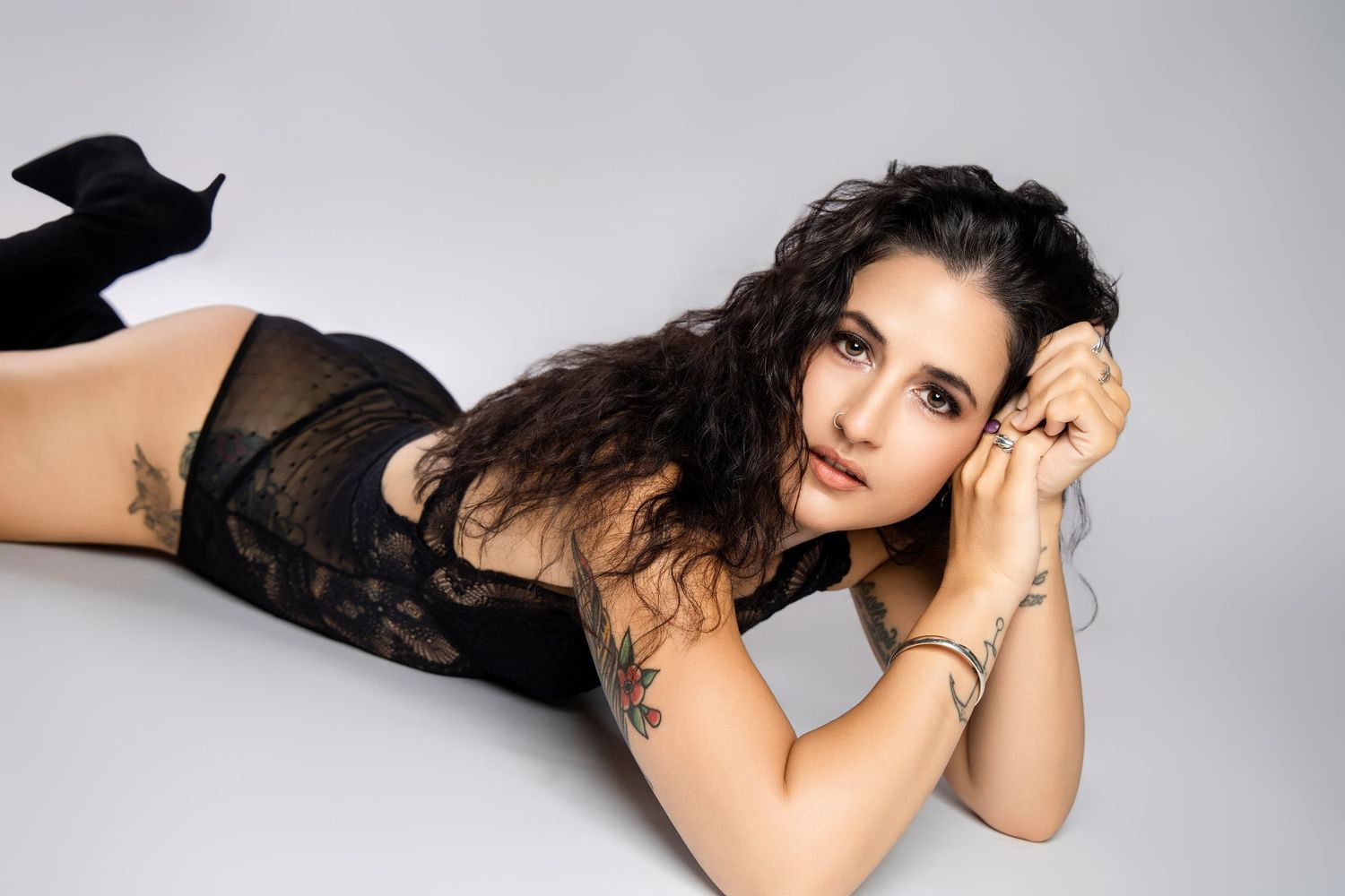 Tattooed brunette woman in black lace bodysuit lying on floor posing for a boudoir style photo shoot.