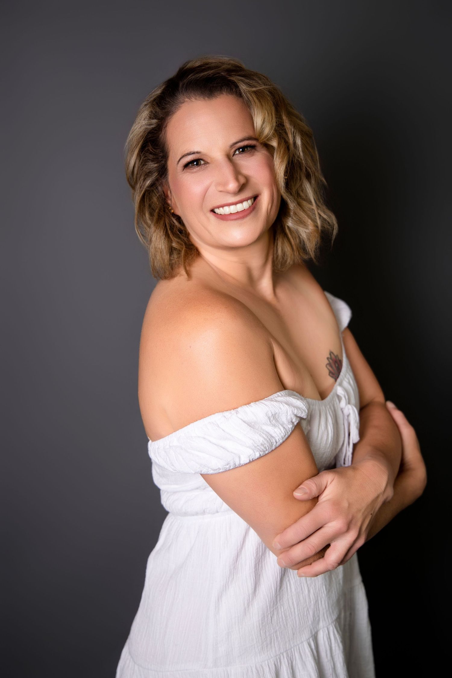 Smiling blonde woman in white off-shoulder dress posing against a dark gray background.