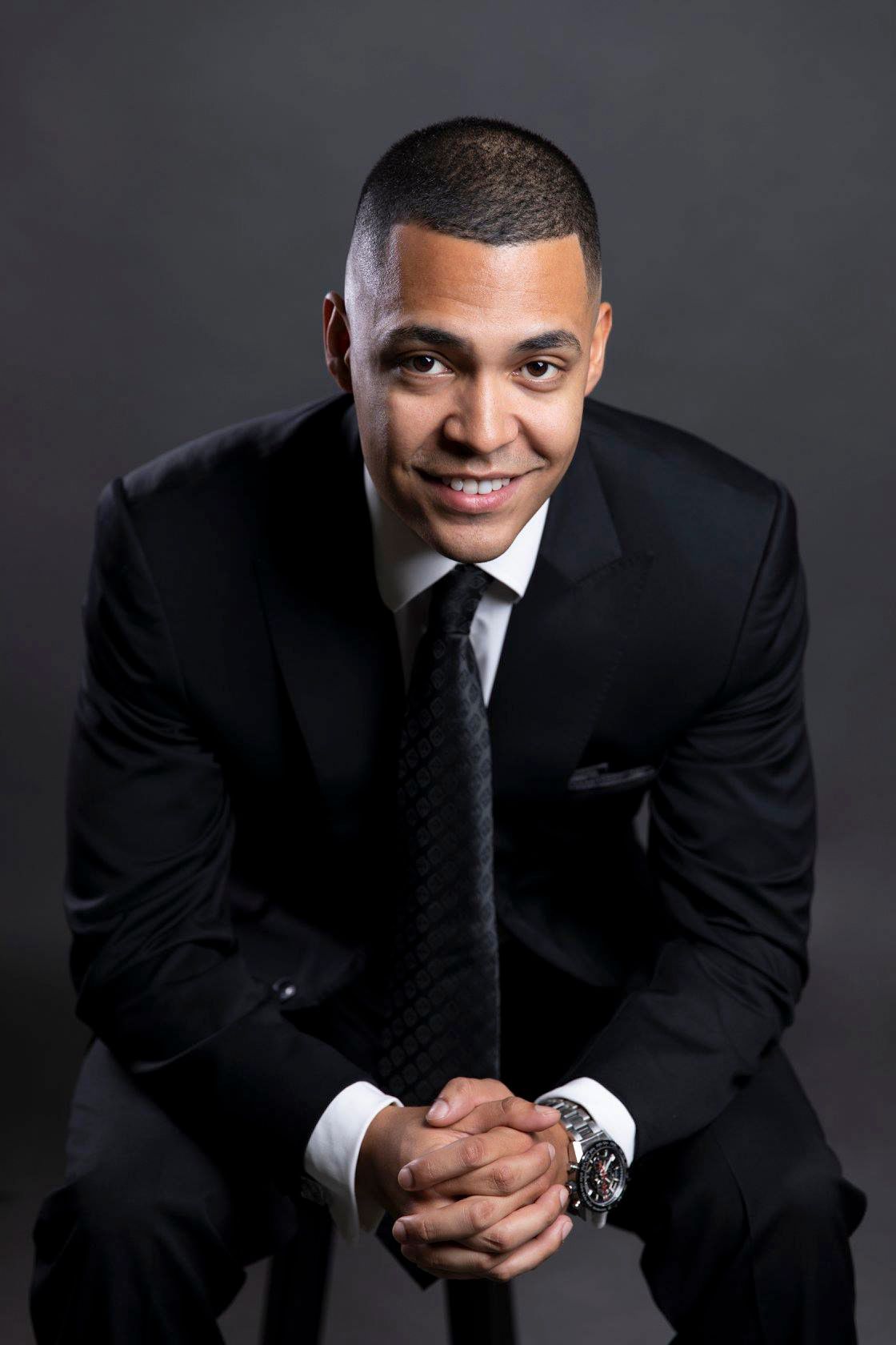 Professional man in black suit and tie sitting confidently against dark studio background.