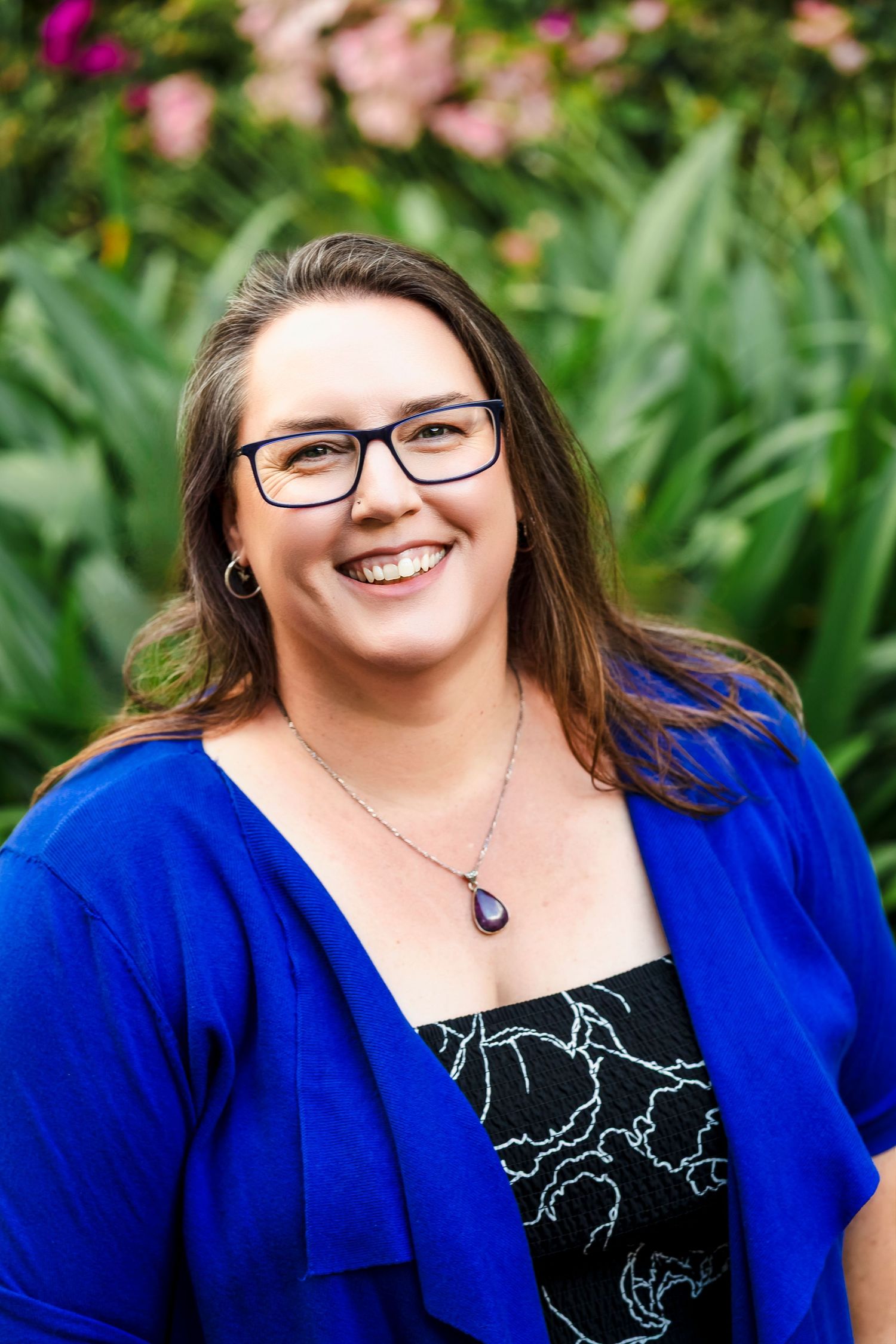 Smiling woman with glasses in blue cardigan poses outdoors with pink flowers in background.
