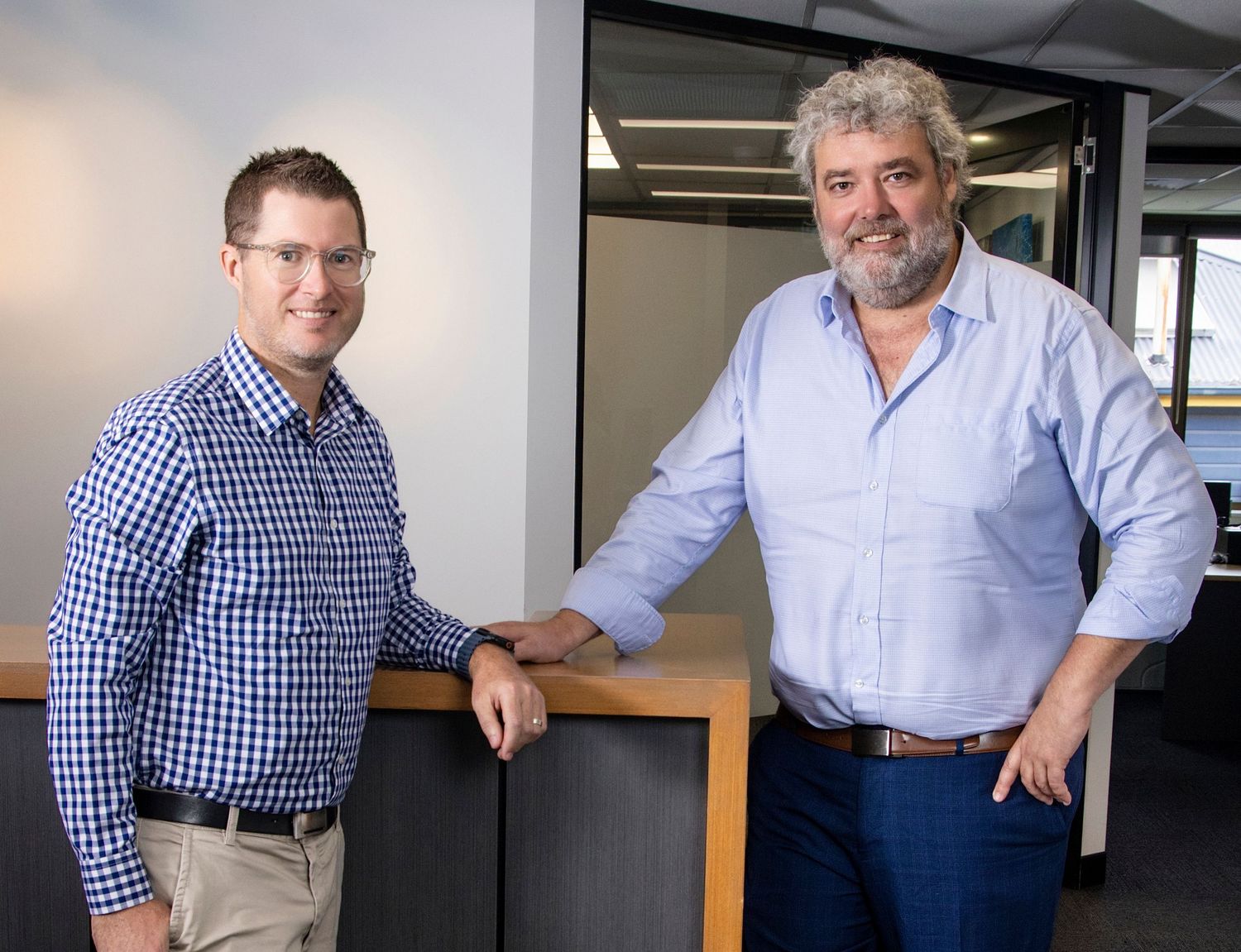 Two professional men smiling and leaning on desk in bright modern office environment.