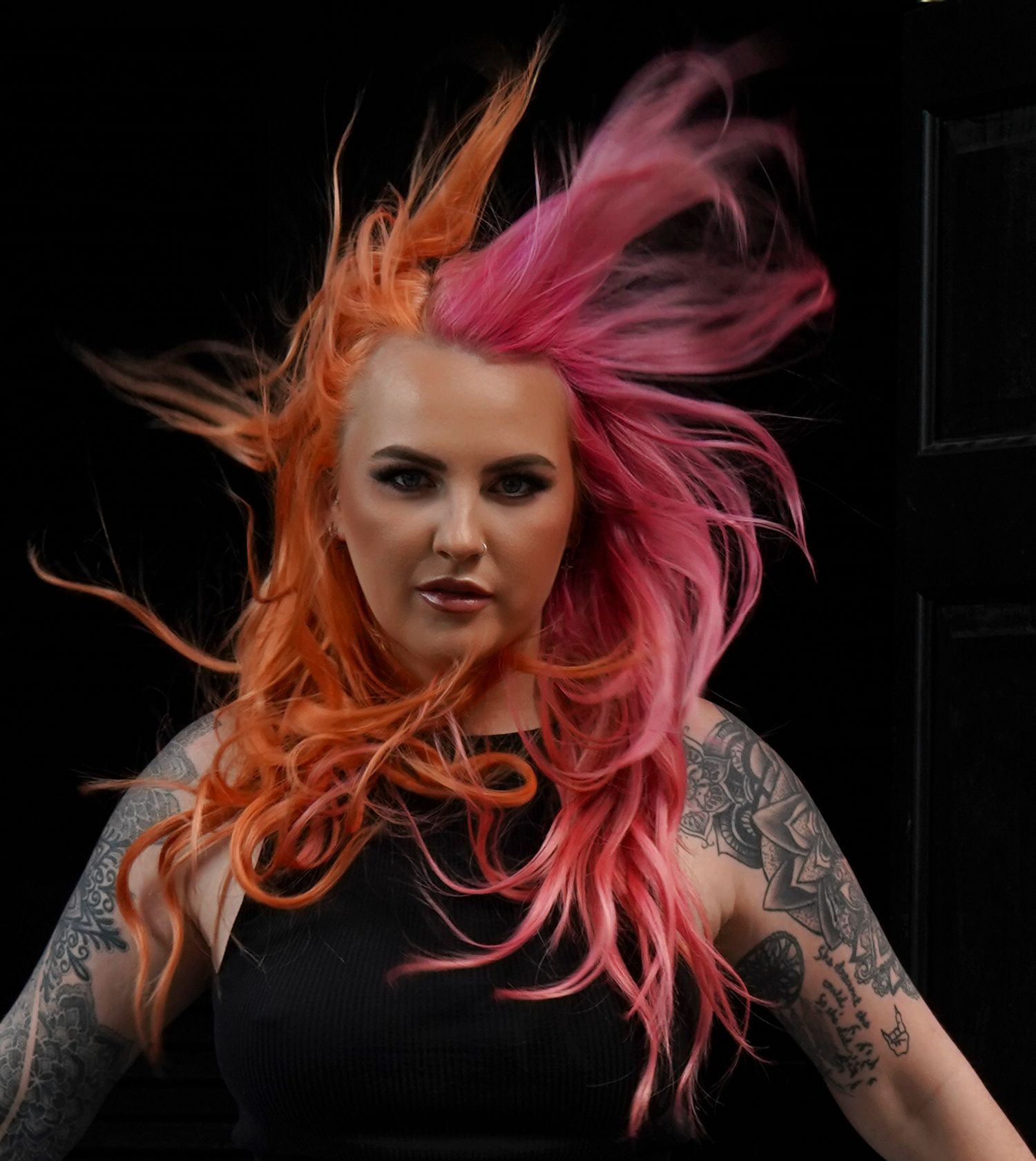 Woman with vibrant orange and pink windswept hair showing arm tattoos outdoors.