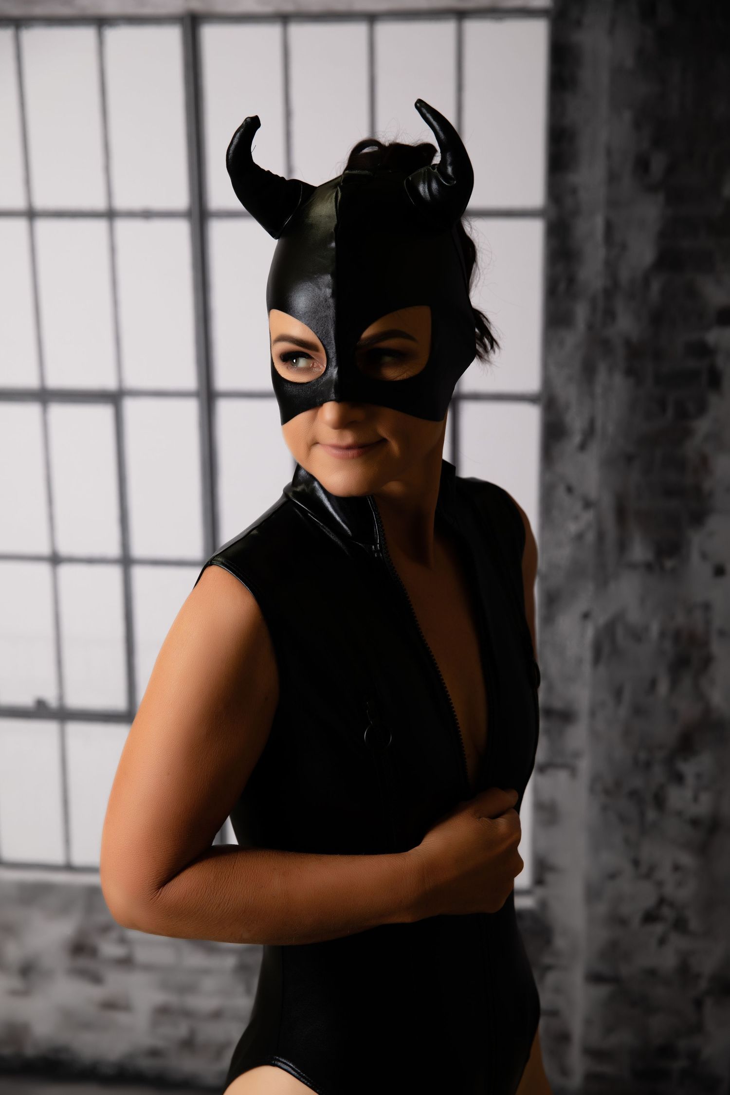 Woman wearing black horned leather mask and black latex bodysuit in industrial style setting.