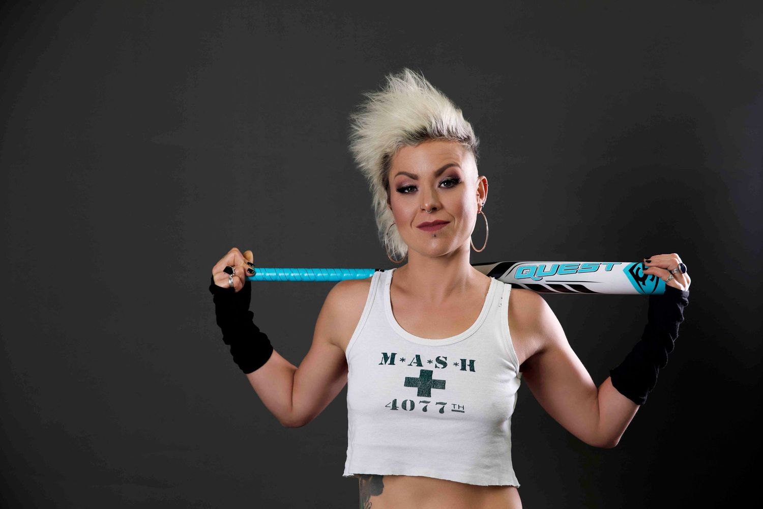 Woman with blonde mohawk wearing white MASH tank top holding blue baseball bat in studio.