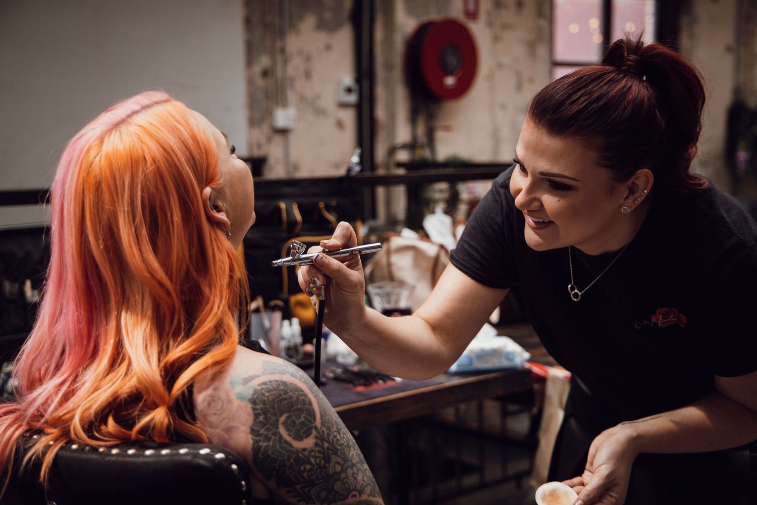 Smiling makeup artist applying airbrush makeup to client with bright pink hair in a professional studio.
