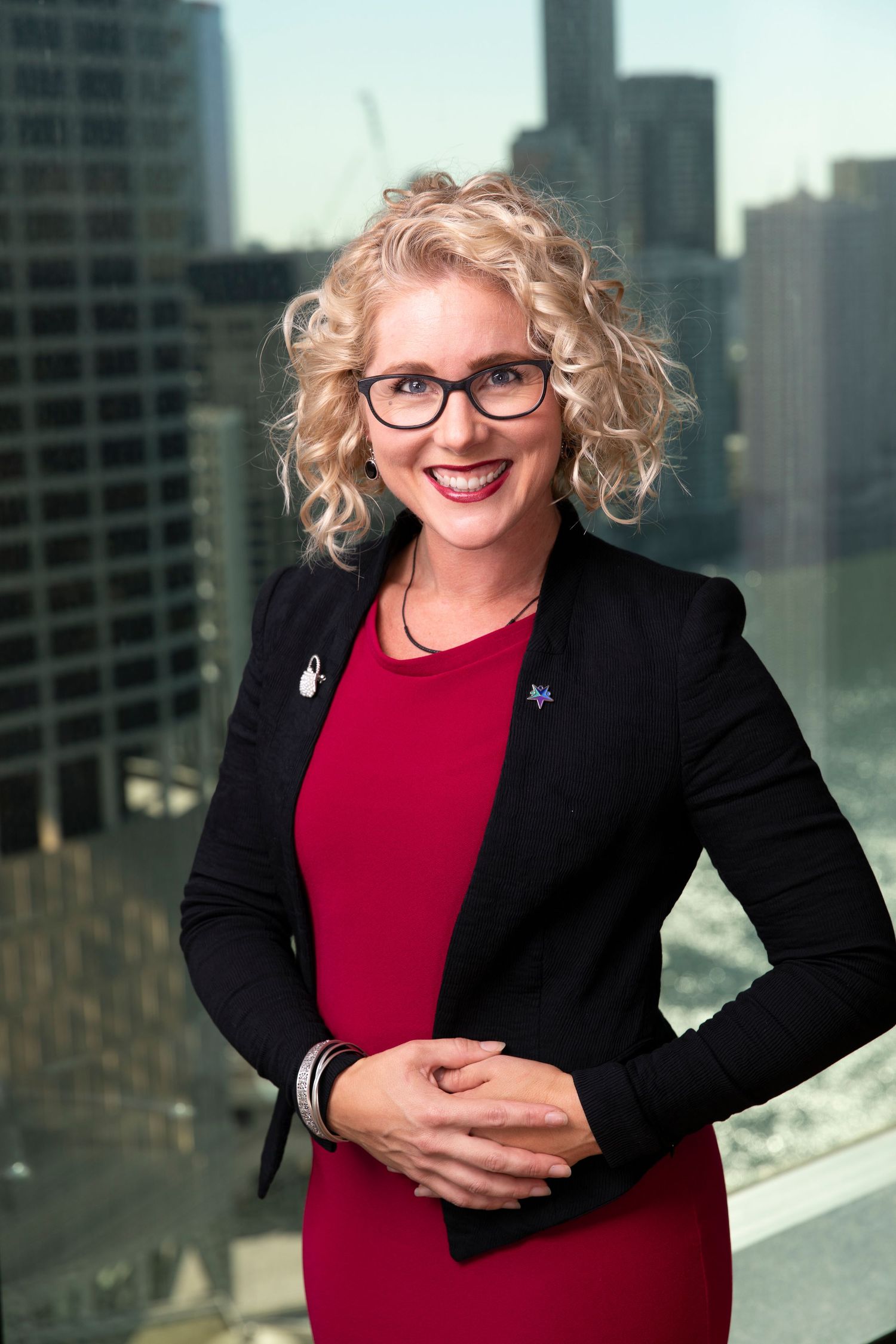 Professional blonde woman in red dress and black blazer smiling with city skyline background.