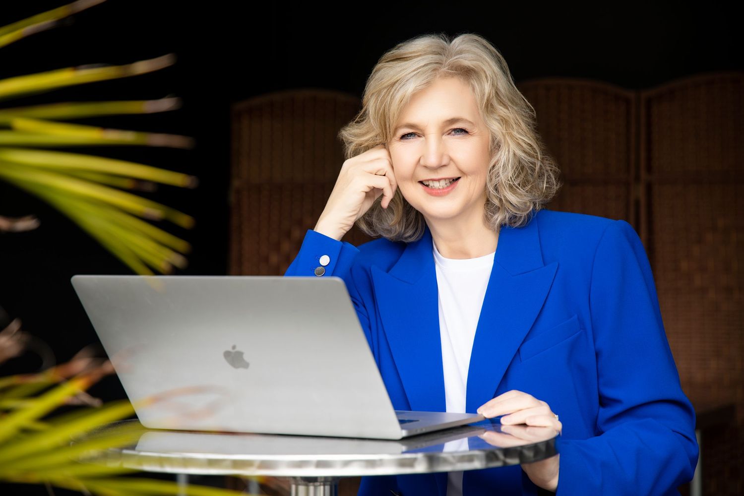 Mature woman with silver hair smiling at laptop wearing blue blazer in outdoor setting.