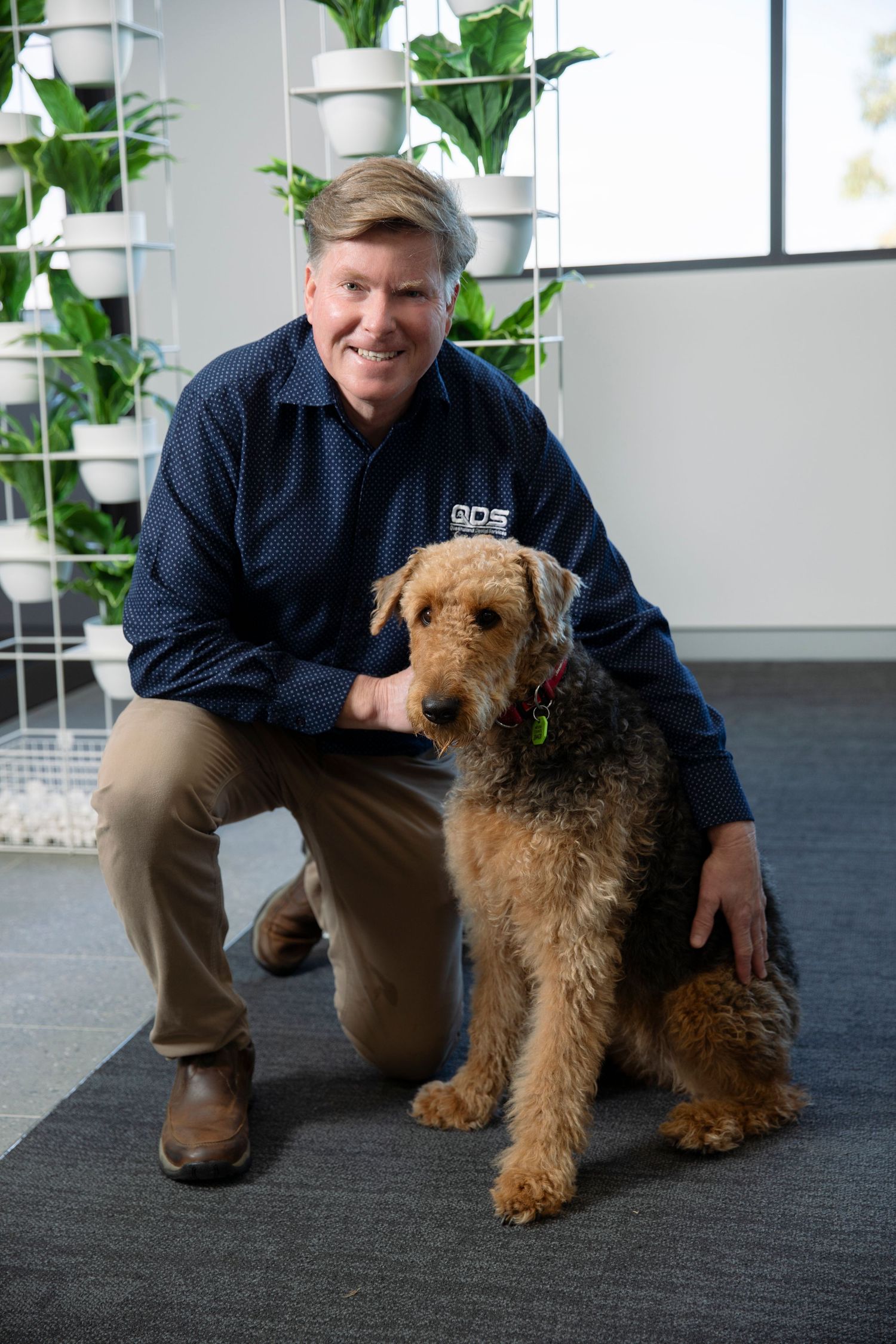 Smiling man in navy shirt kneeling beside an Airedale Terrier dog in a modern office.
