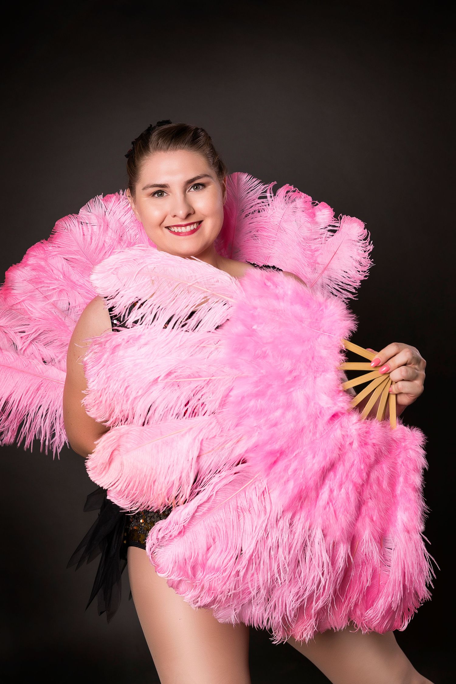 Woman in burlesque costume poses with large pink ostrich feather fans.