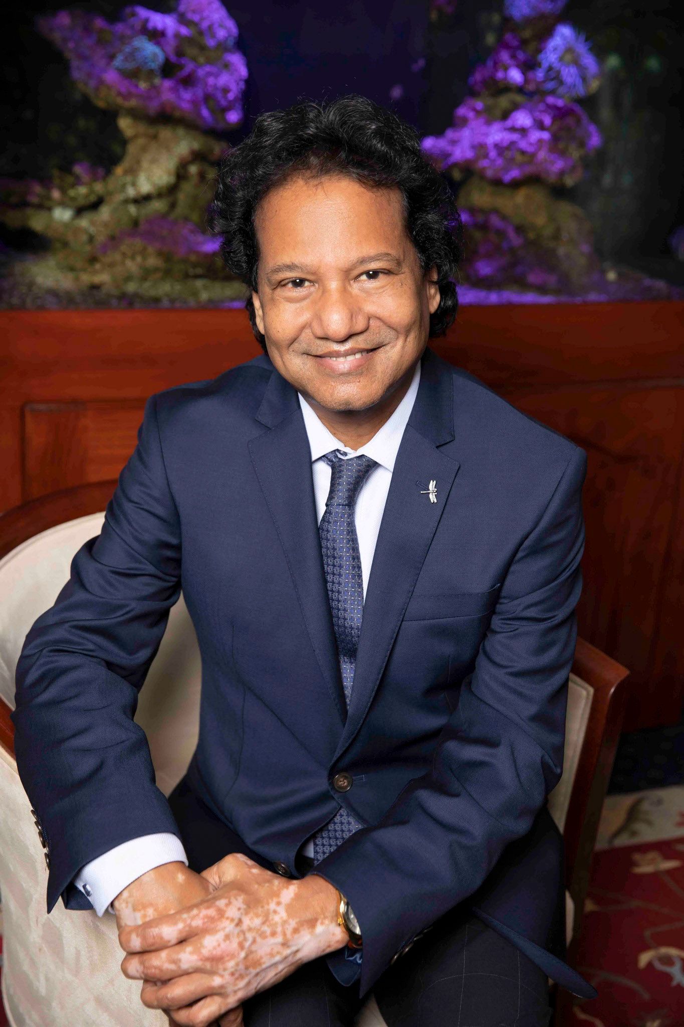 Smiling man in navy blue suit seated in elegant room with illuminated aquarium background.