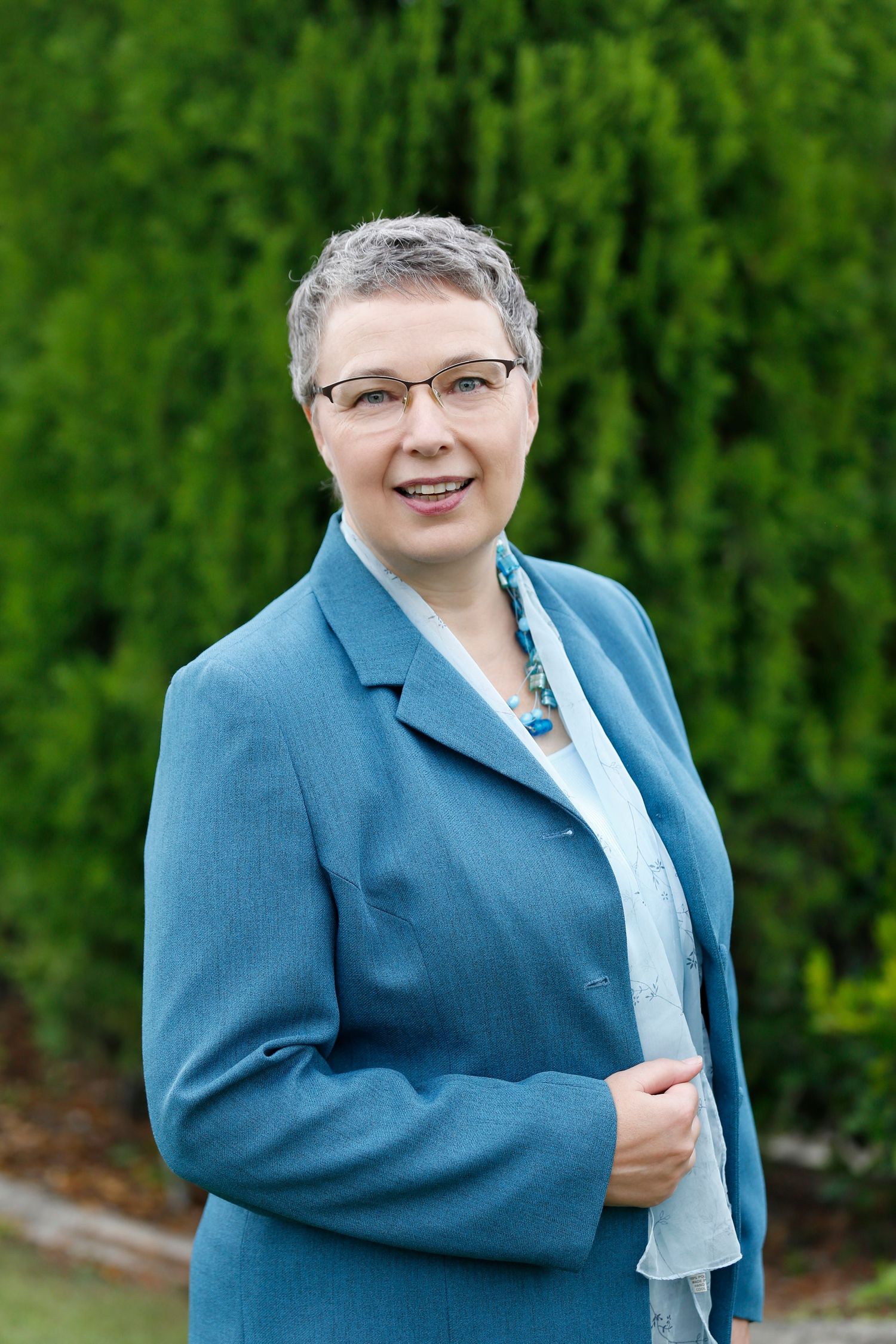Professional woman with short gray hair wearing blue blazer posing outdoors near green hedges.