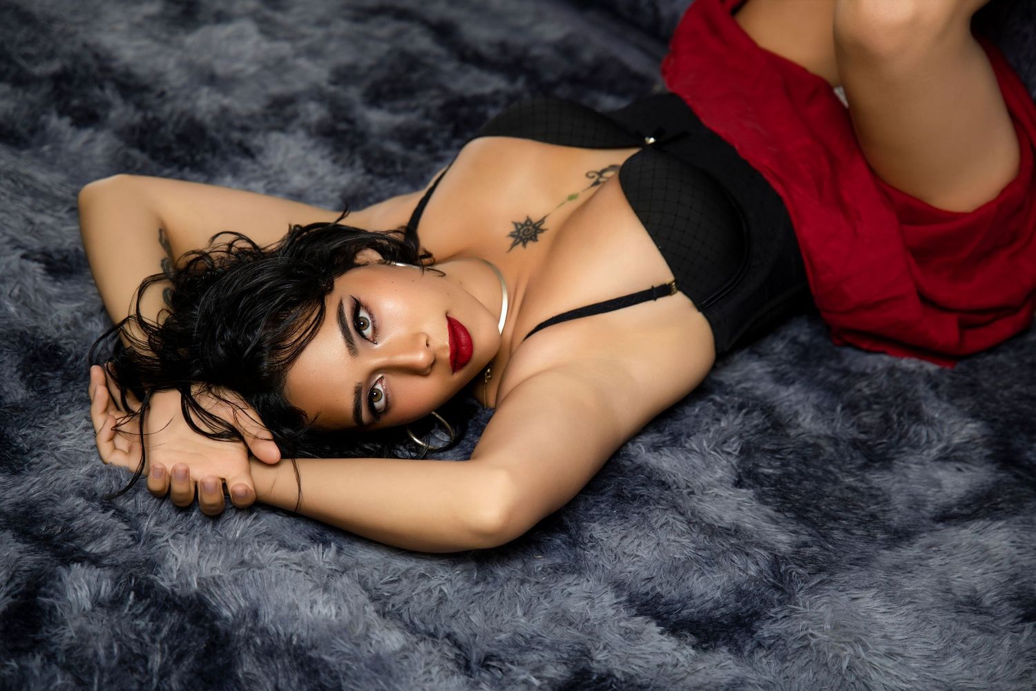Sultry brunette woman in black lingerie with red lips lying on gray fur rug in boudoir photo.