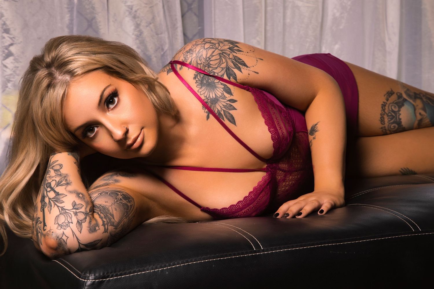 Tattooed blonde woman in red lace lingerie posing seductively on dark leather surface.