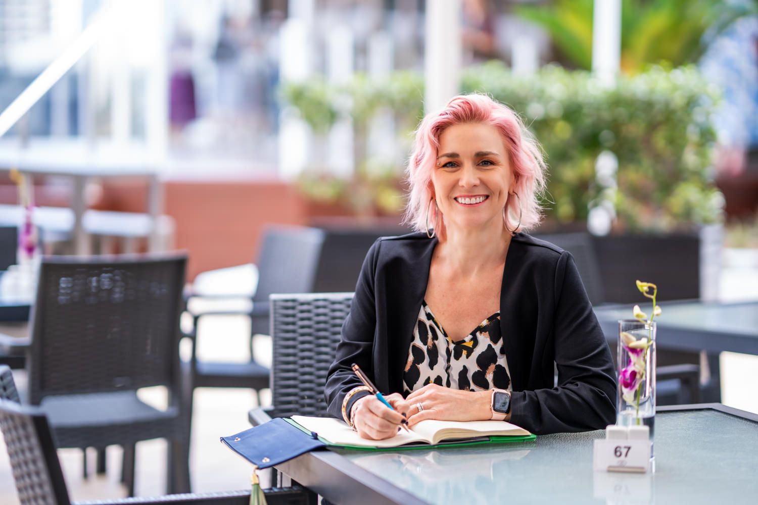 Woman with pink hair writing in notebook at outdoor cafe table, smiling.