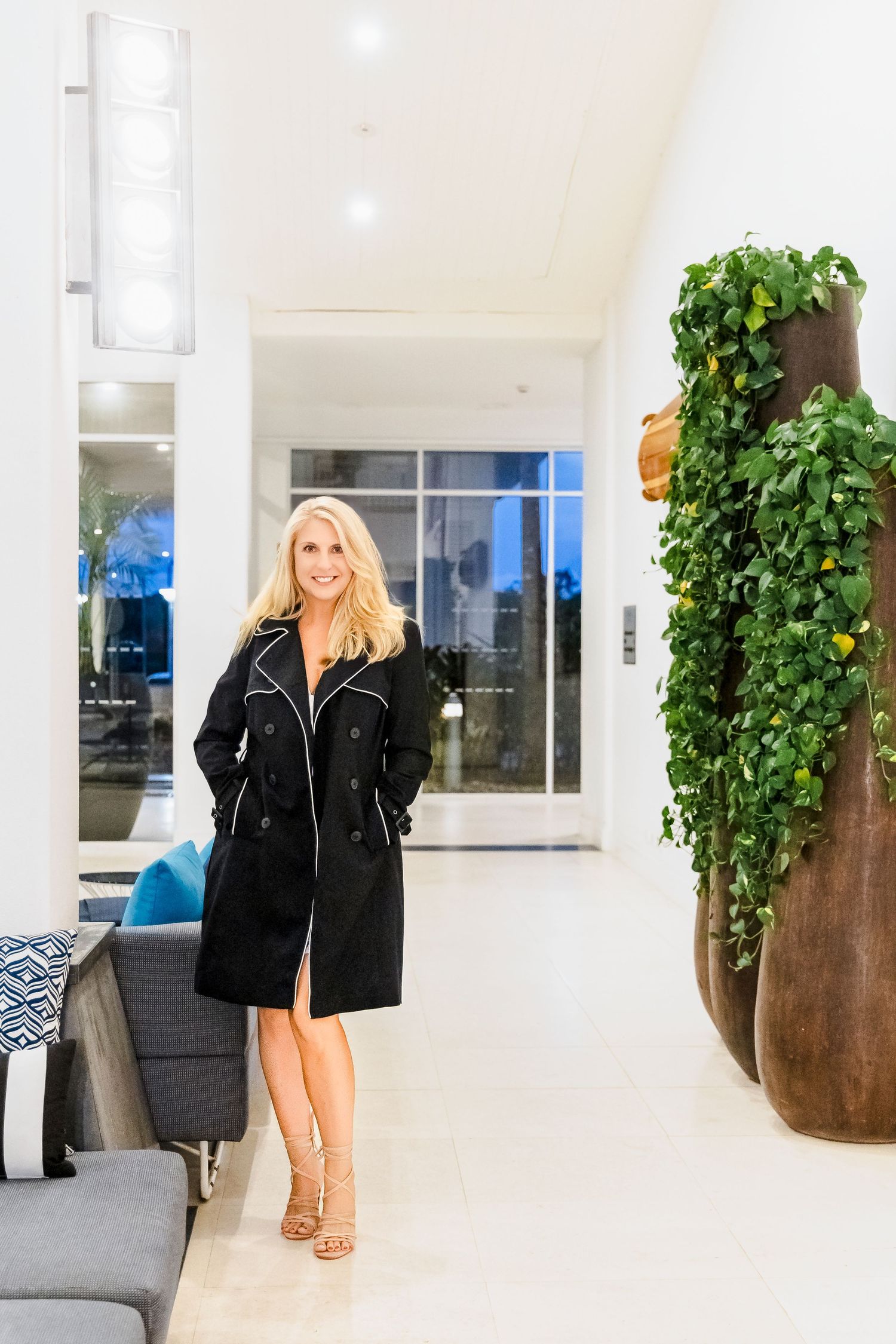 Blonde woman in black trench coat standing in a modern white hotel lobby interior.