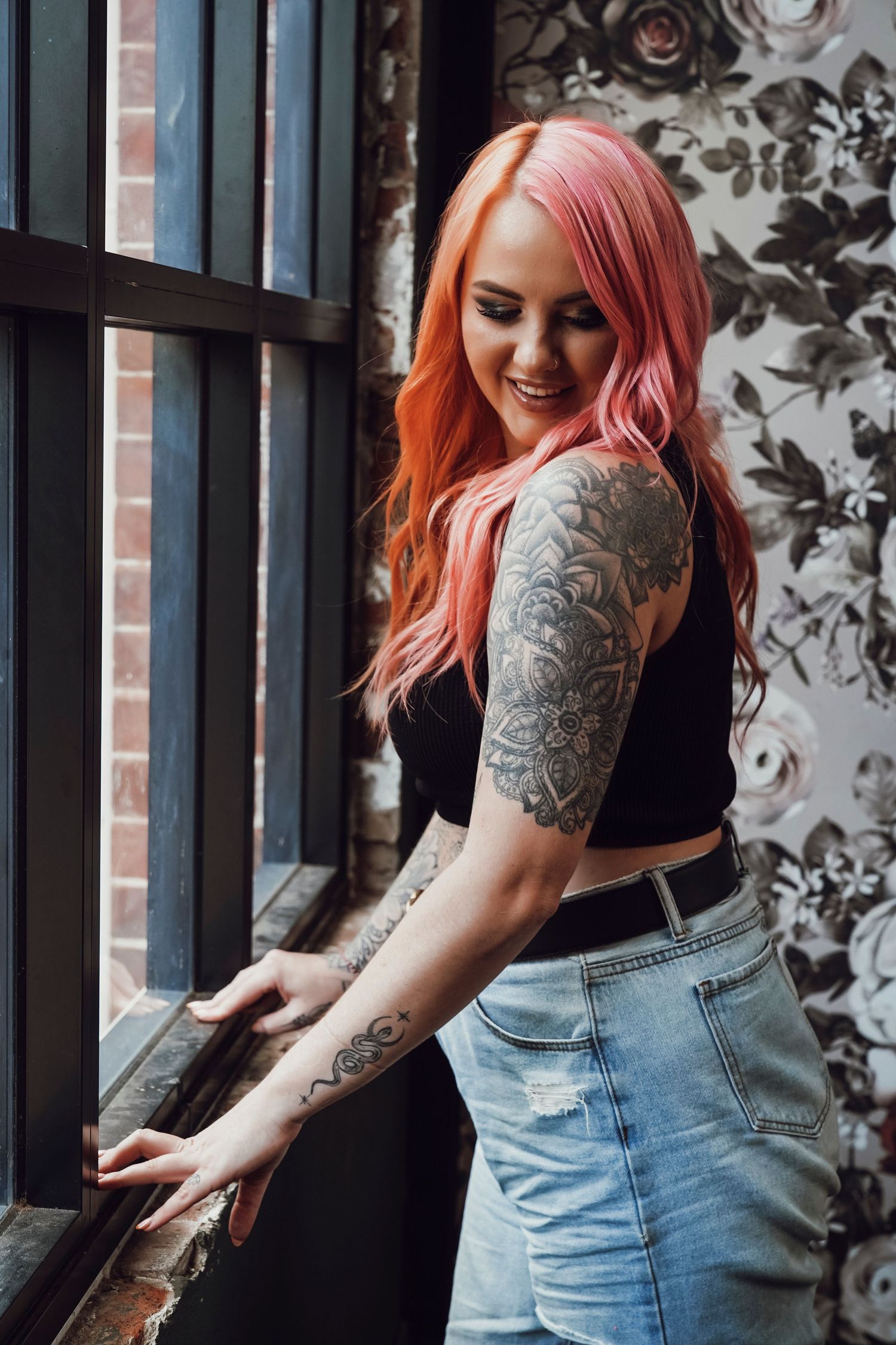 Tattooed woman with pink hair smiles by window against floral wallpaper in jeans.