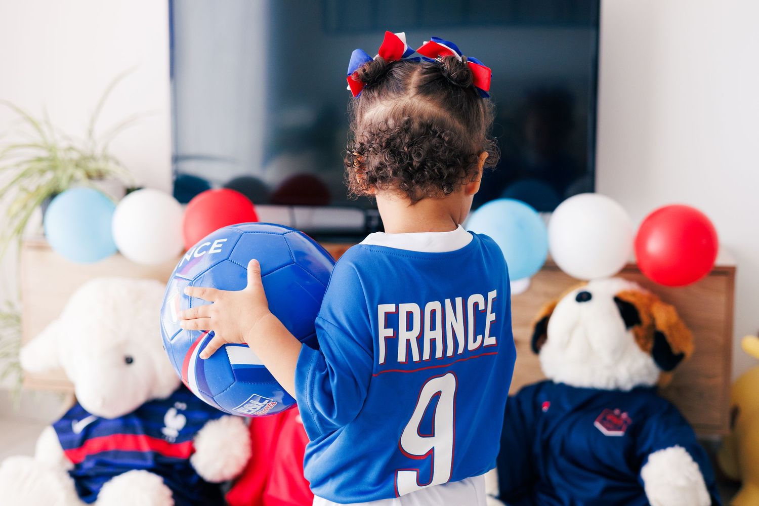 Une petite fille en tenue de l’équipe de France tient un ballon de football devant une décoration bleu, blanc et rouge.