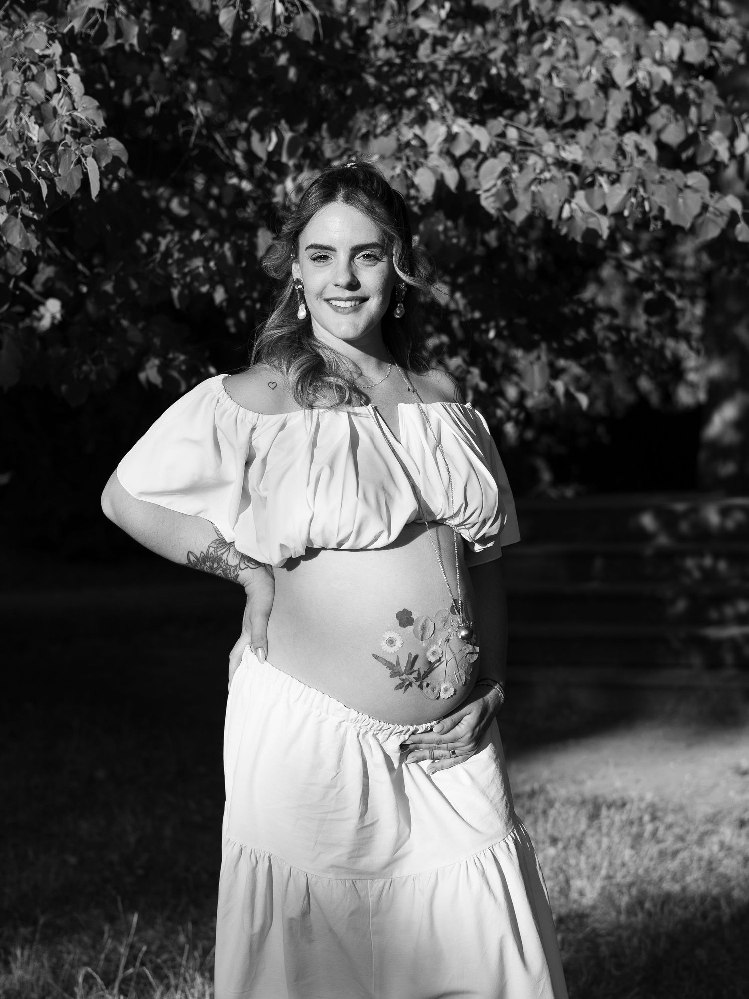 Une future maman pose en noir et blanc en tenant son ventre fleuri pendant une séance photo grossesse en extérieur.