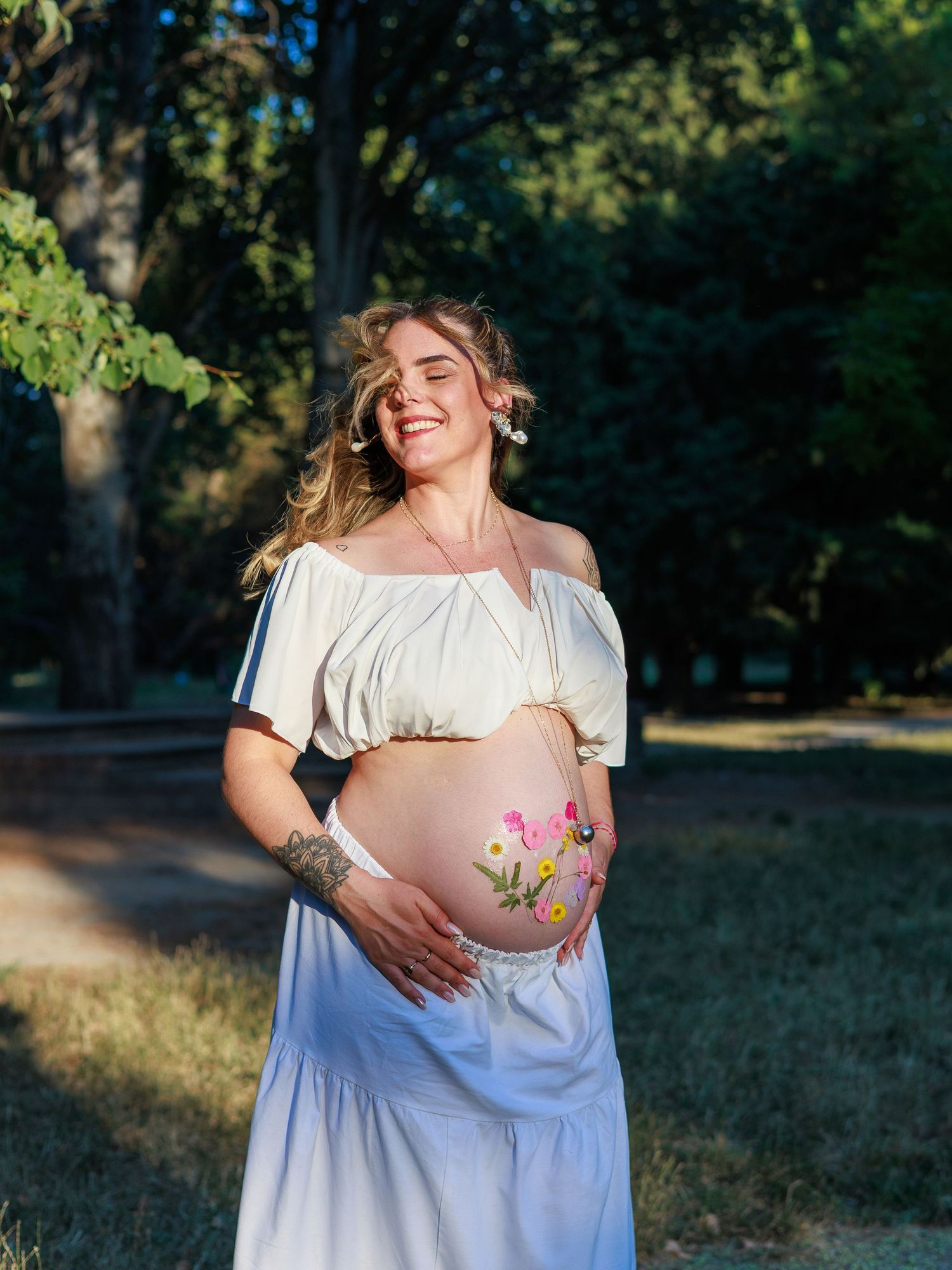 Une future maman sourit en tenant son ventre pendant une séance photo grossesse en extérieur.