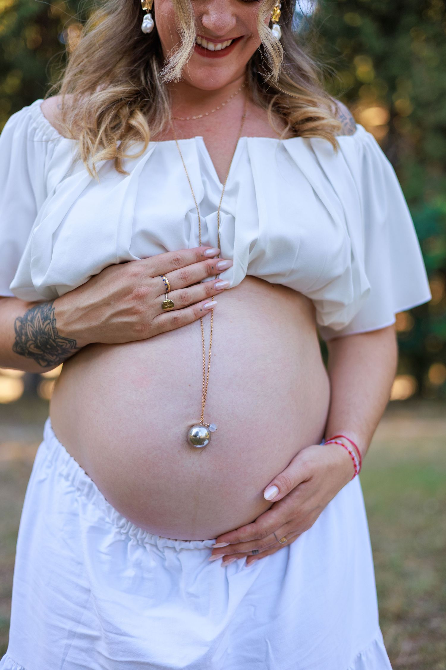 Une future maman sourit en tenant son ventre pendant une séance photo grossesse en extérieur.
