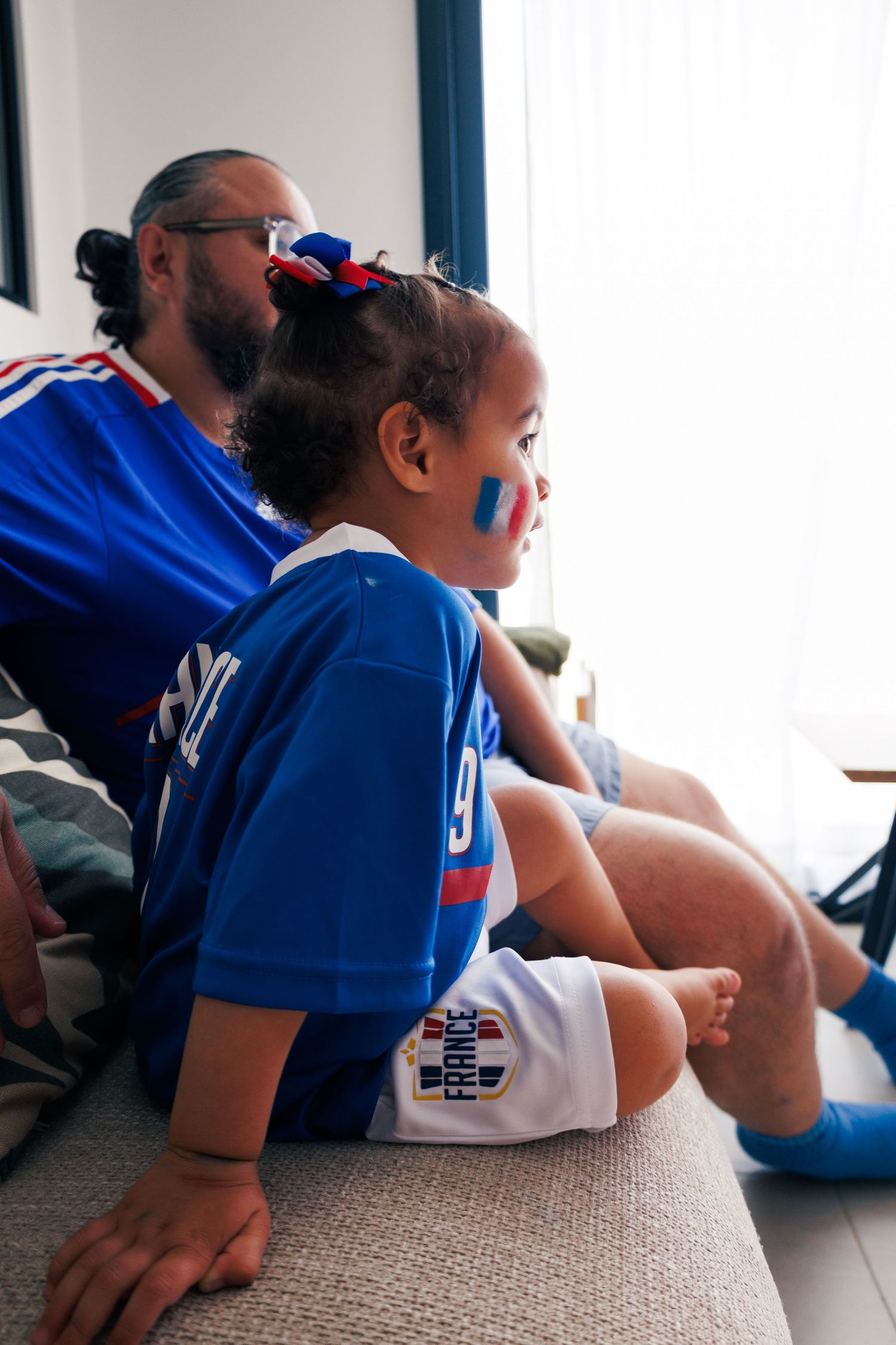Une petite fille regarde un match de football avec son père, tous les deux habillés aux couleurs de l’équipe de France.