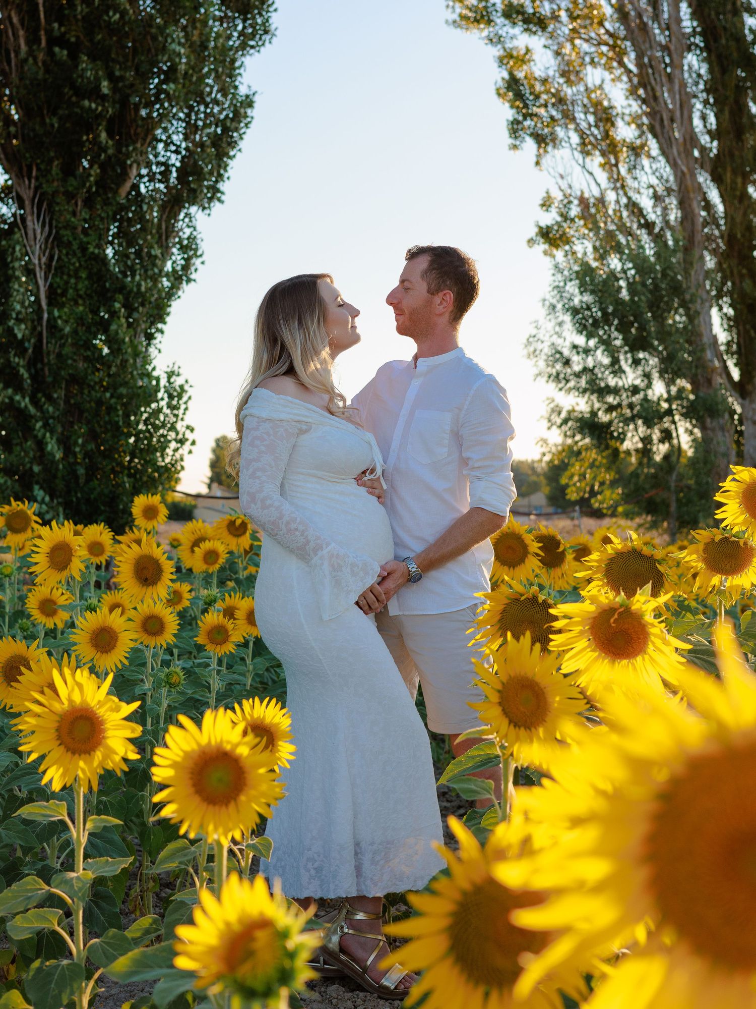 Un couple de futurs parents se tient dans un champ de tournesols pendant une séance photo grossesse en extérieur.