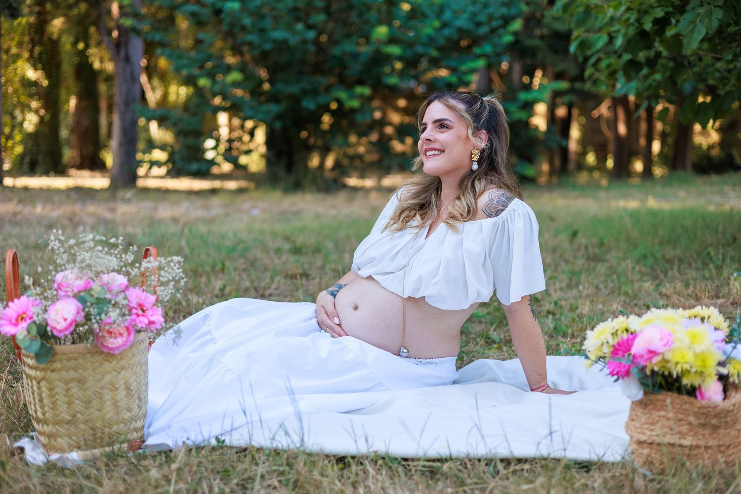 Une future maman est assise dans l’herbe entourée de fleurs pendant une séance photo grossesse en extérieur.