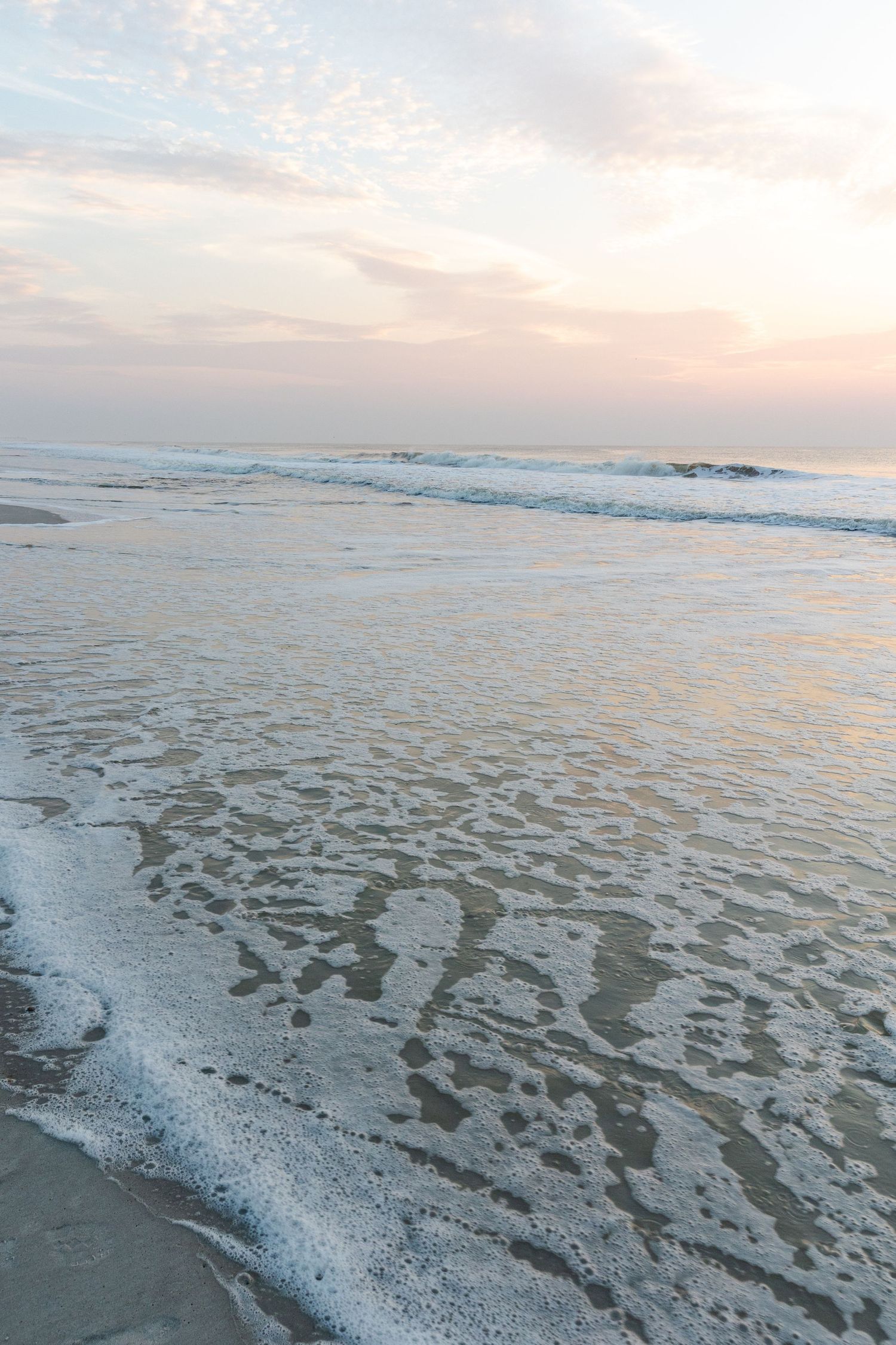 Gentle ocean waves wash up on a sandy beach at sunset with soft pink clouds in the pastel sky.