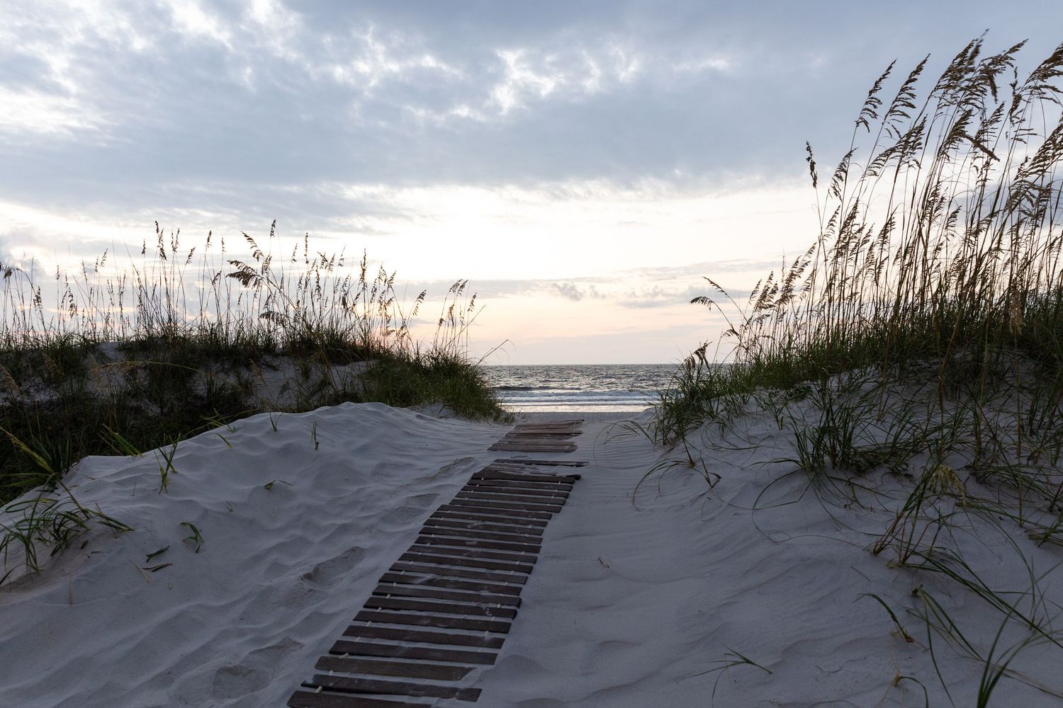 Wooden boardwalk path through sand dunes with sea oats leads to ocean beach under cloudy sunset sky at dusk.