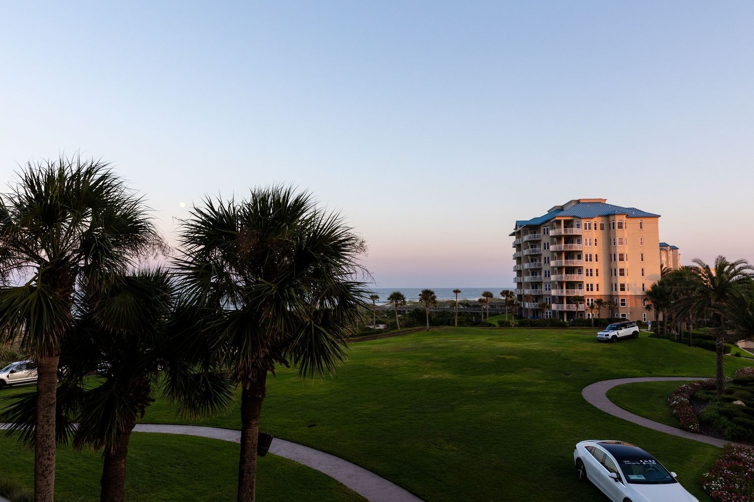 Palm trees and a curved walkway lead to a beachfront condo building at sunset in Florida.