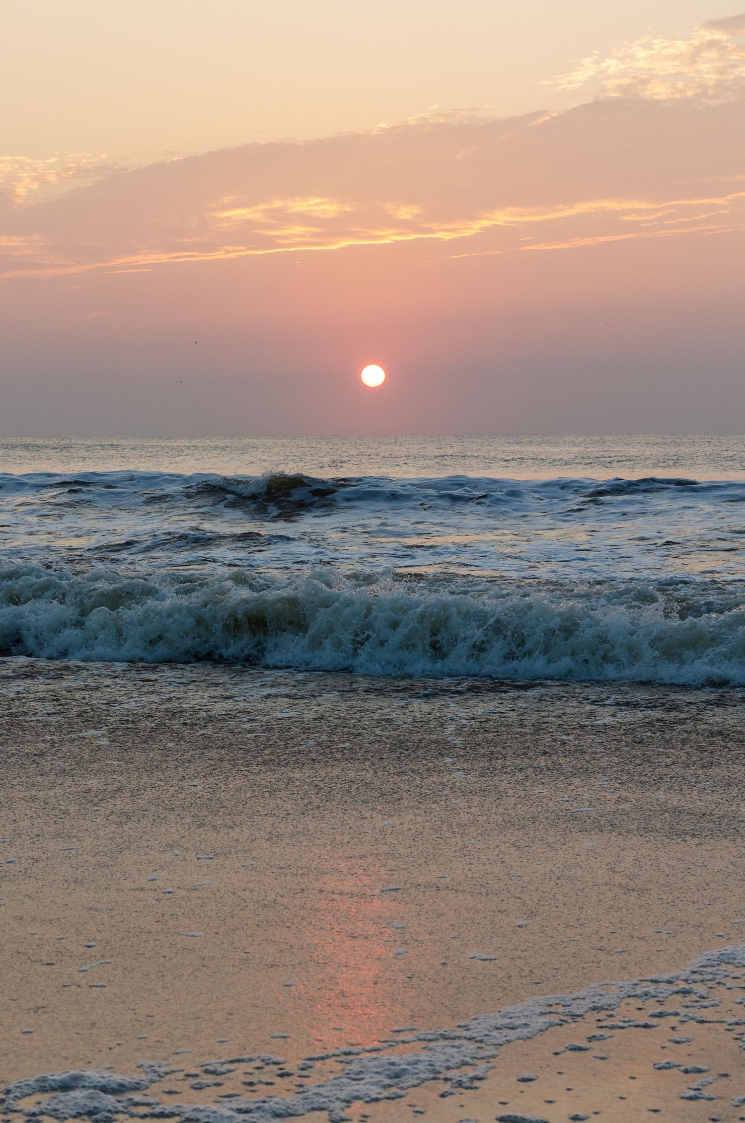 Ocean waves crash on the beach during a stunning pink sunset as the sun dips below the horizon over calm waters.