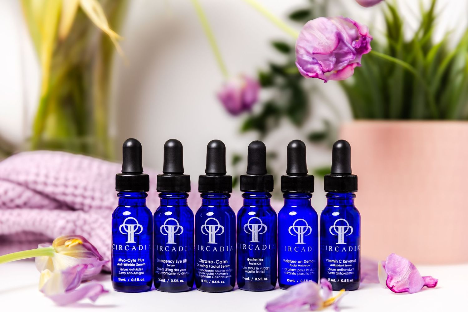 Row of six blue glass dropper bottles with matching labels displayed with purple flowers.