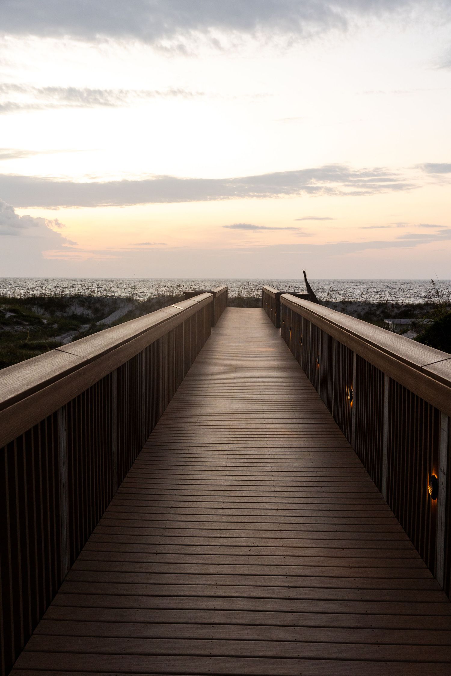 A wooden boardwalk pathway stretches towards the horizon at sunset with cloudy skies and ocean views.