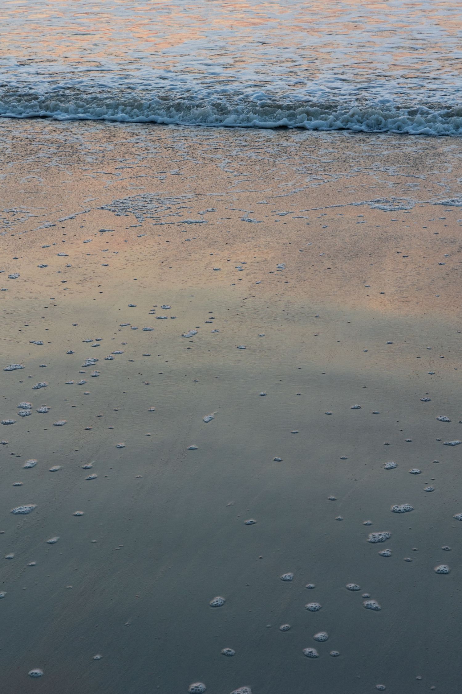 Gentle waves wash over sandy beach at sunset creating reflective patterns and small bubbles on wet shoreline.
