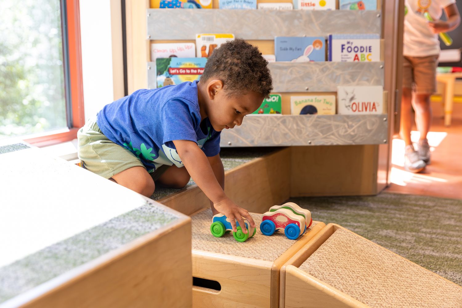 A young child plays with toy cars on wooden blocks in a sunny playroom.